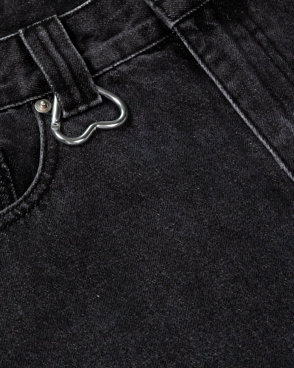 Detailaufnahme der Gürtelschlaufe mit silbernem Herz-Karabiner auf schwarzem Denim. Die grobe Stoffstruktur und Nahtverarbeitung sind sichtbar.