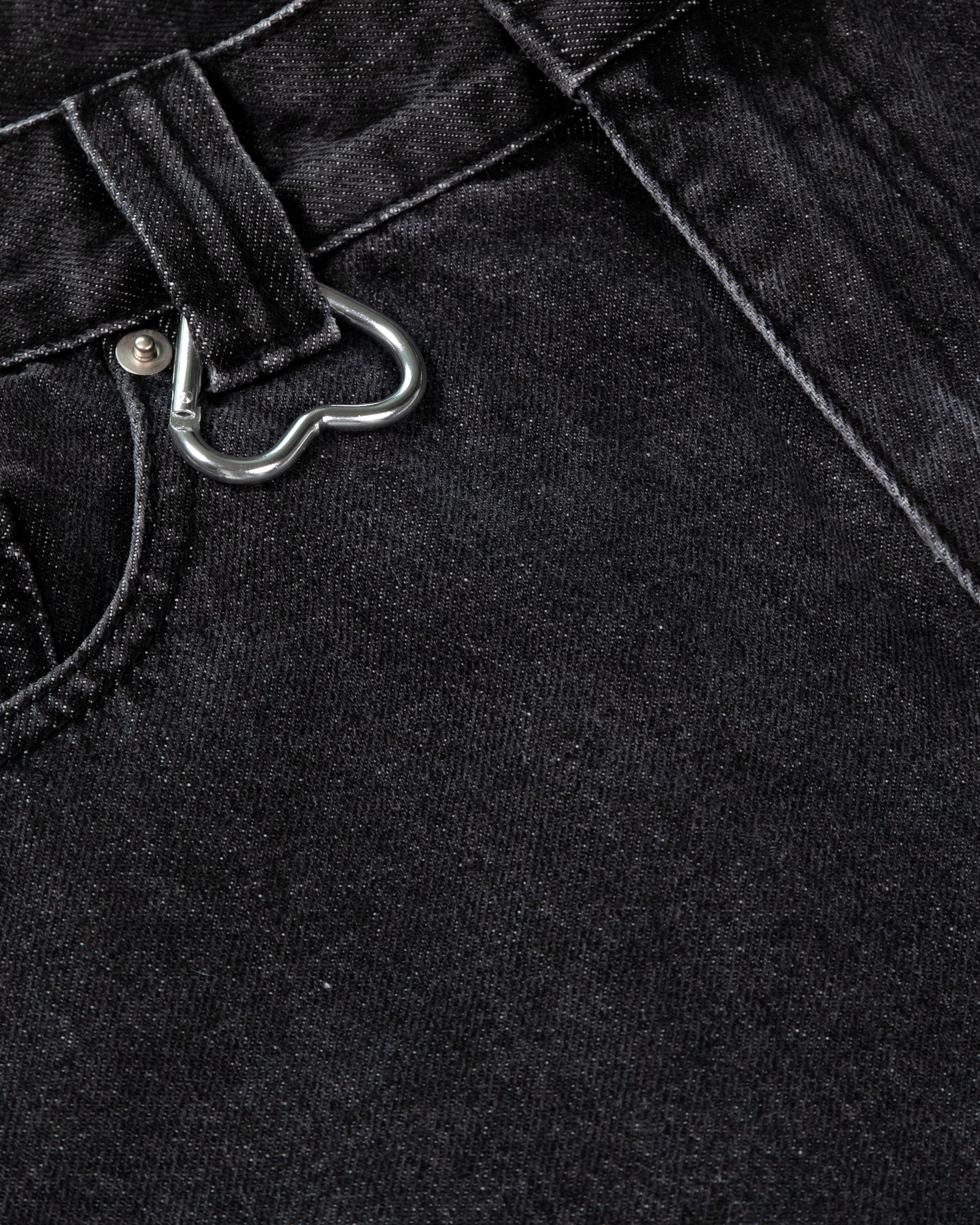 Detailaufnahme der Gürtelschlaufe mit silbernem Herz-Karabiner auf schwarzem Denim. Die grobe Stoffstruktur und Nahtverarbeitung sind sichtbar.