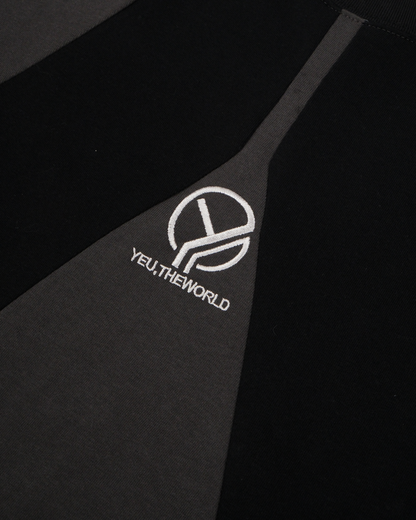 Person trägt ein zweifarbiges T-Shirt mit grafisch geschwungenen Farbflächen in Schwarz auf graugrünem Grund und zentraler Logo-Stickerei „YEU.THEWORLD“ auf der Brust.