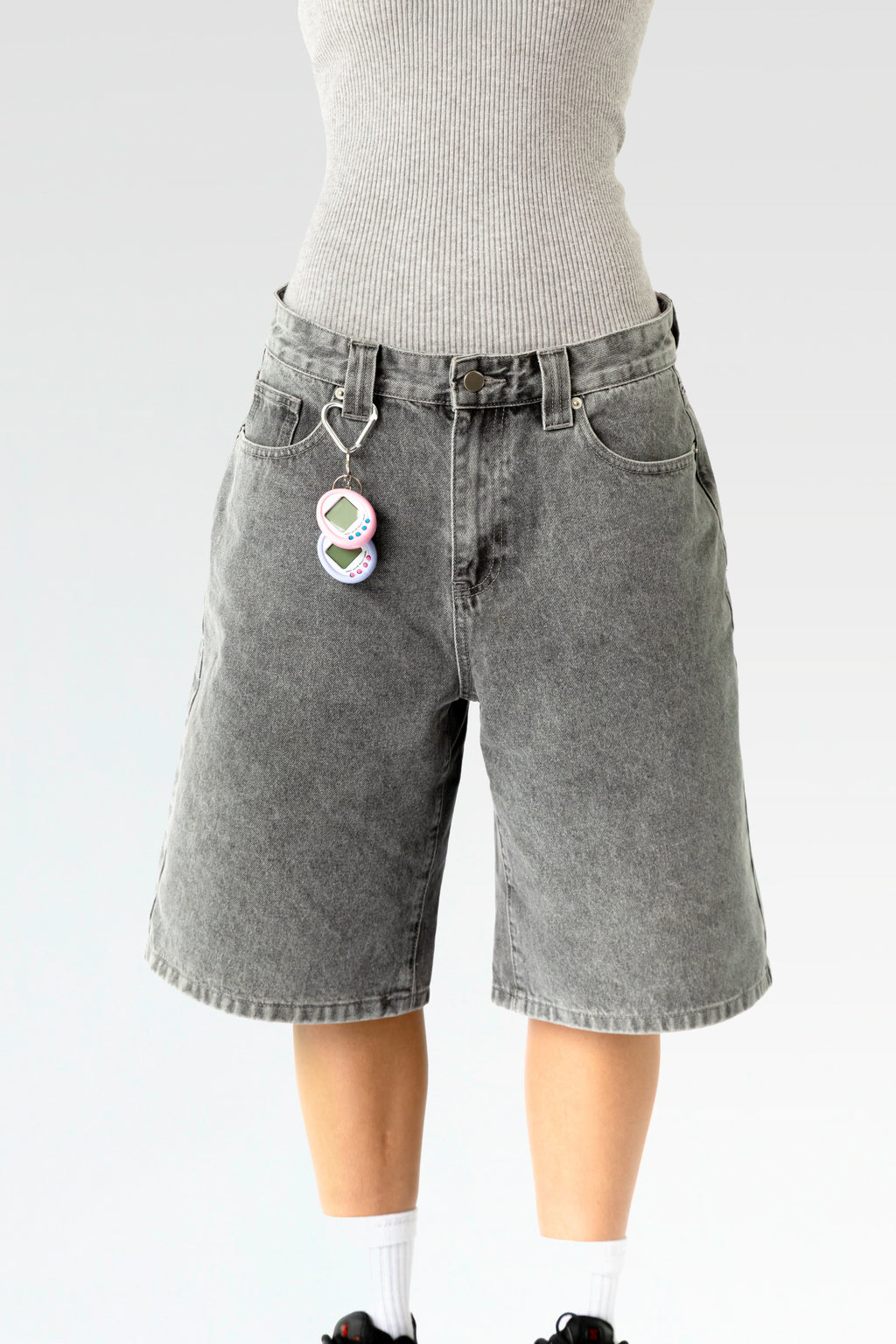 Vorderansicht einer hellgrau verwaschenen, knielangen Jeansshorts aus Waschdenim an einer stehenden Person. Die Shorts hat einen Herz-förmigen Karabiner an einer Gürtelschlaufe, daran hängt ein pink-weißes elektronisches Tamagotchi-Spielzeug. Sichtbar sind der klassische 5-Pocket-Schnitt, Gürtelschlaufen und ein Metallknopf am Bund.
