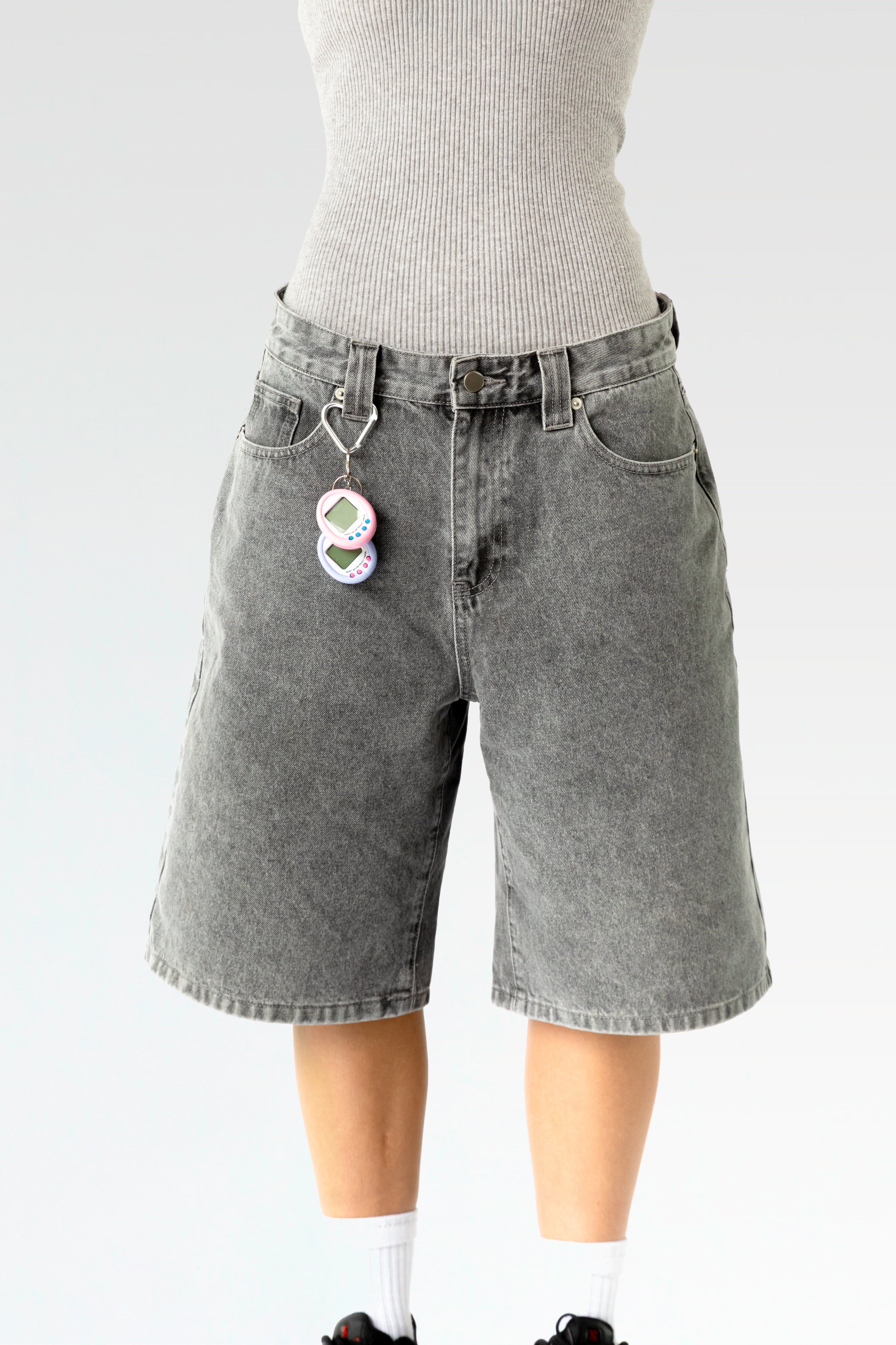 Vorderansicht einer hellgrau verwaschenen, knielangen Jeansshorts aus Waschdenim an einer stehenden Person. Die Shorts hat einen Herz-förmigen Karabiner an einer Gürtelschlaufe, daran hängt ein pink-weißes elektronisches Tamagotchi-Spielzeug. Sichtbar sind der klassische 5-Pocket-Schnitt, Gürtelschlaufen und ein Metallknopf am Bund.