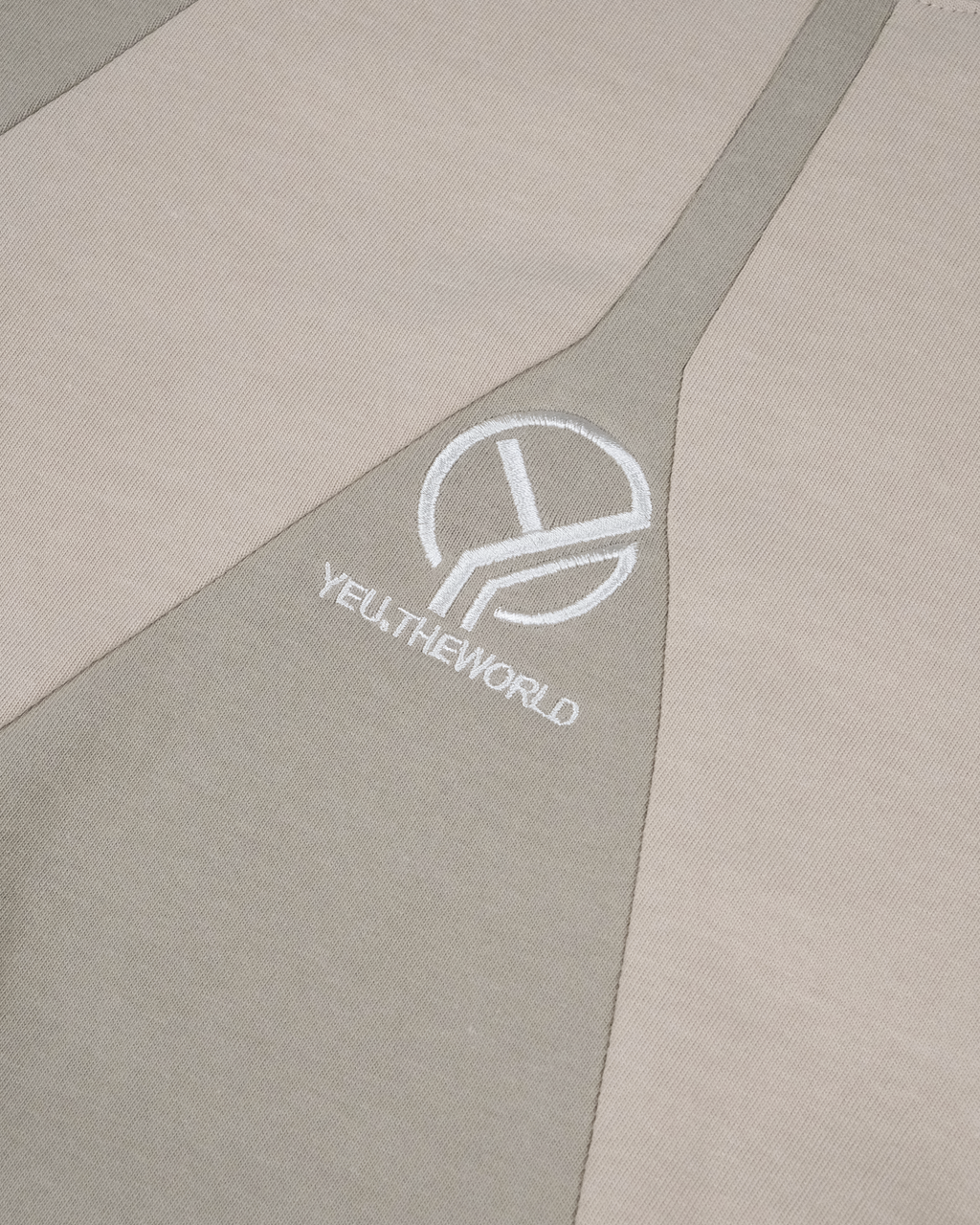 Detailansicht eines cremefarbenen T-Shirts mit hellbeigem Einsatz in V-Form. Im Zentrum des Einsatzes ist das weiße Logo „YEU.THEWORLD“ hochwertig eingestickt.
