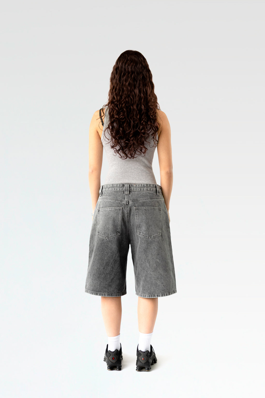 Rückansicht einer hellgrau verwaschenen, weit geschnittenen Jeansshorts aus gewaschenem Denim. Die Shorts reicht bis knapp übers Knie und zeigt zwei aufgesetzte Gesäßtaschen. Getragen von einer stehenden Person mit langen Locken, sportlichen Sneakers und weißem Tanktop.