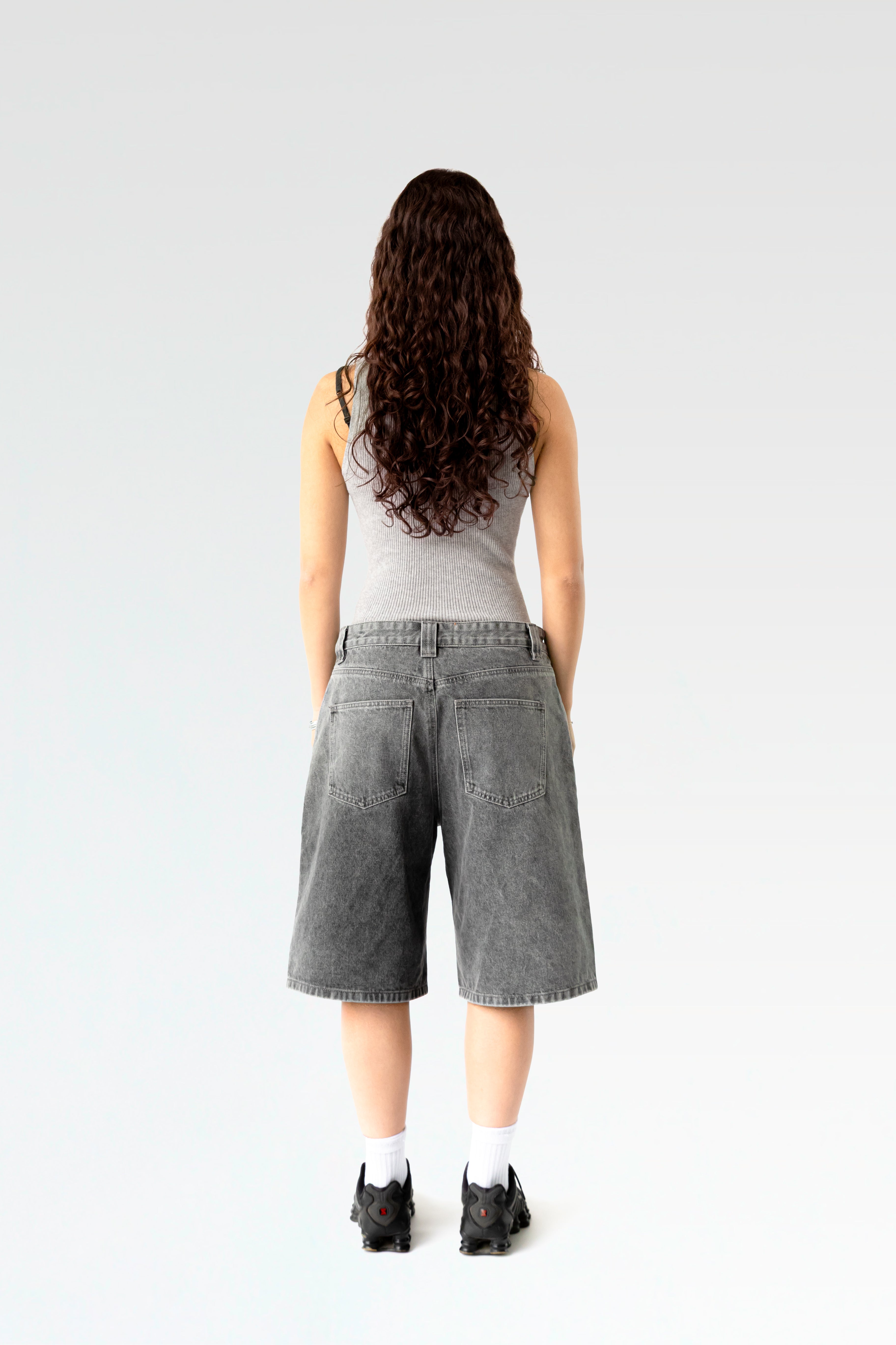 Rückansicht einer hellgrau verwaschenen, weit geschnittenen Jeansshorts aus gewaschenem Denim. Die Shorts reicht bis knapp übers Knie und zeigt zwei aufgesetzte Gesäßtaschen. Getragen von einer stehenden Person mit langen Locken, sportlichen Sneakers und weißem Tanktop.