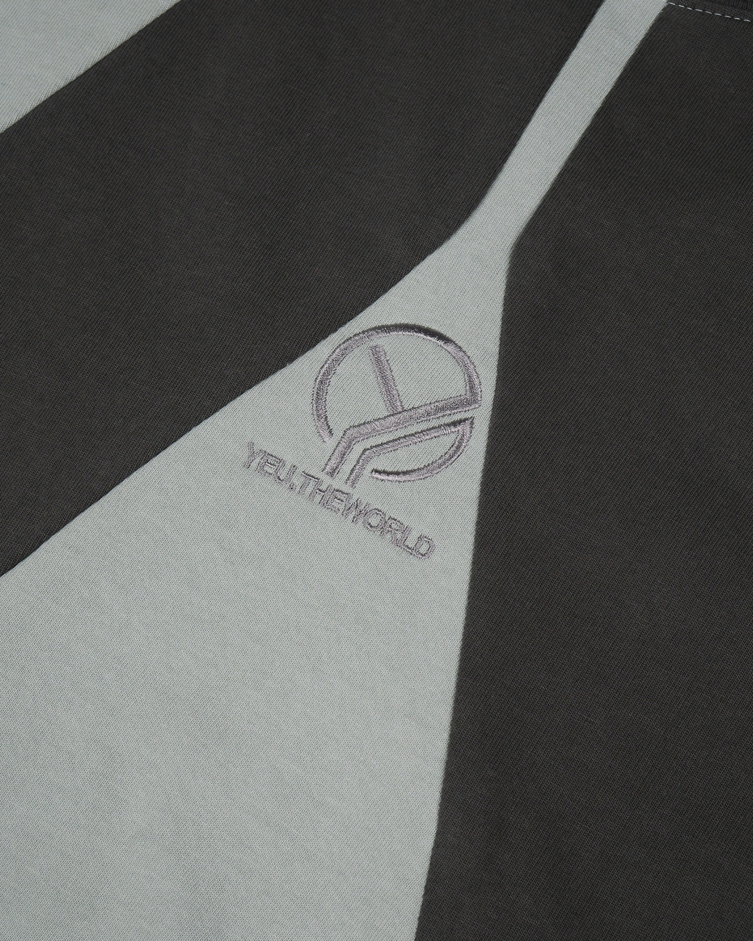 Nahansicht eines zweifarbigen T-Shirts in dunklem Graugrün und Schwarz mit mittig platzierter, fein gestickter „YEU.THEWORLD“-Wort-Bild-Marke in Grau.