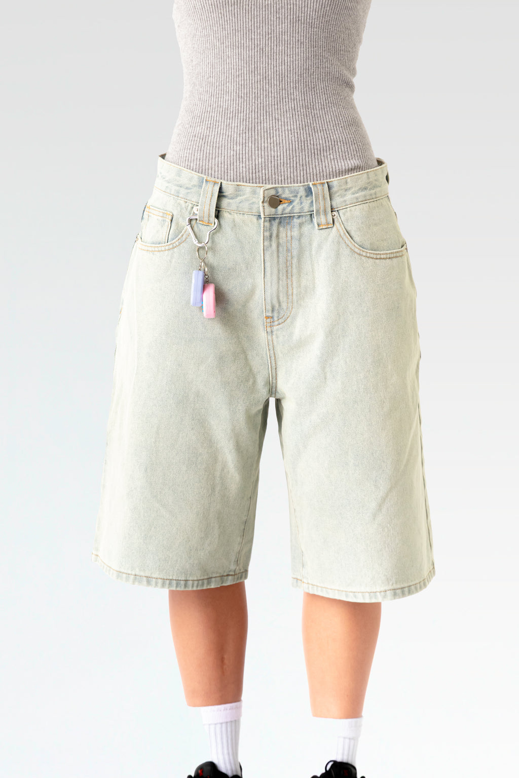 Vorderansicht einer hellblauen, knielangen Jeansshorts aus Waschdenim an einer stehenden Person. Die Shorts hat einen Herz-förmigen Karabiner an einer Gürtelschlaufe, daran hängt ein pink-weißes elektronisches Tamagotchi-Spielzeug. Sichtbar sind der klassische 5-Pocket-Schnitt, Gürtelschlaufen und ein Metallknopf am Bund.
