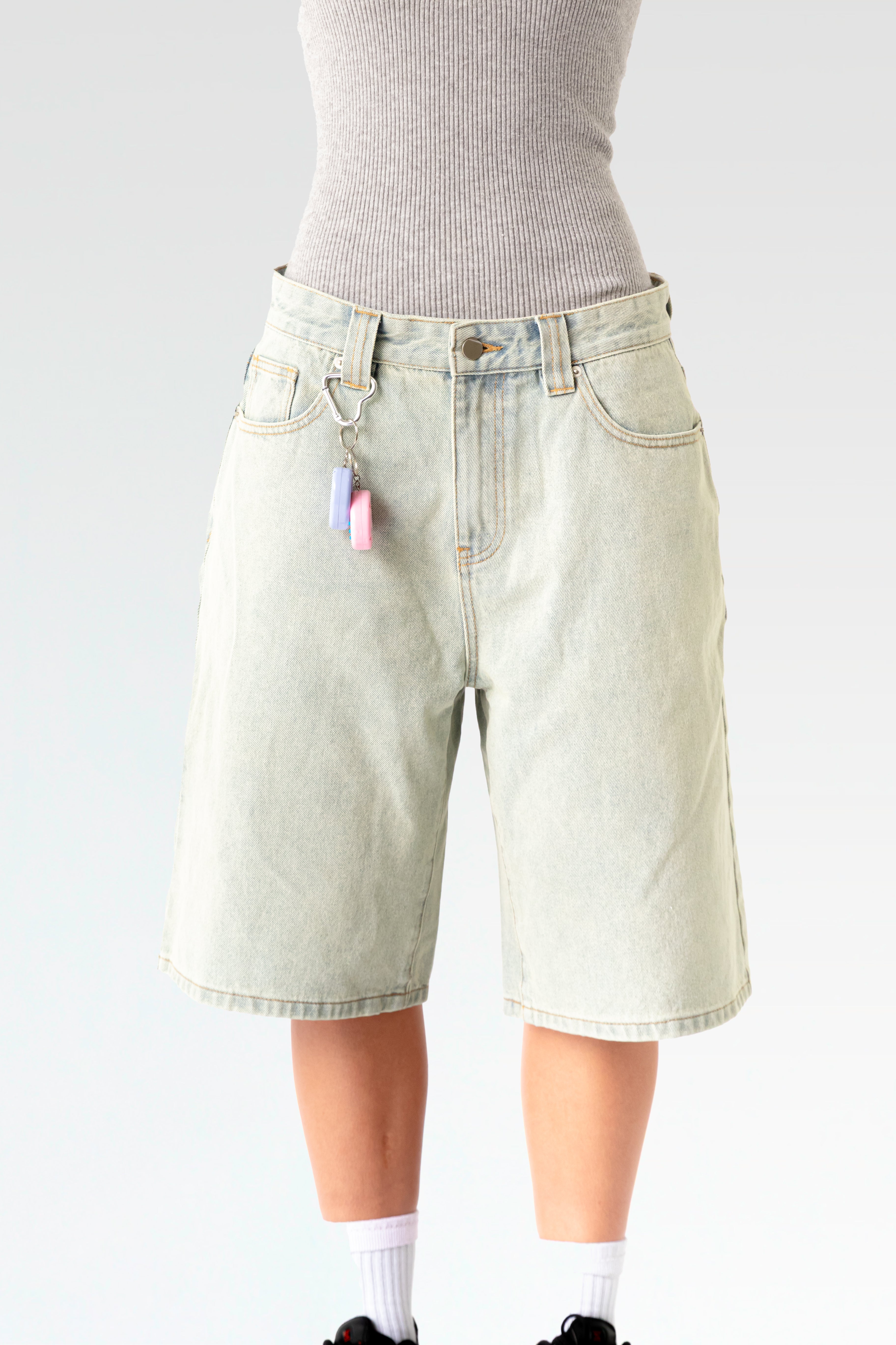 Vorderansicht einer hellblauen, knielangen Jeansshorts aus Waschdenim an einer stehenden Person. Die Shorts hat einen Herz-förmigen Karabiner an einer Gürtelschlaufe, daran hängt ein pink-weißes elektronisches Tamagotchi-Spielzeug. Sichtbar sind der klassische 5-Pocket-Schnitt, Gürtelschlaufen und ein Metallknopf am Bund.
