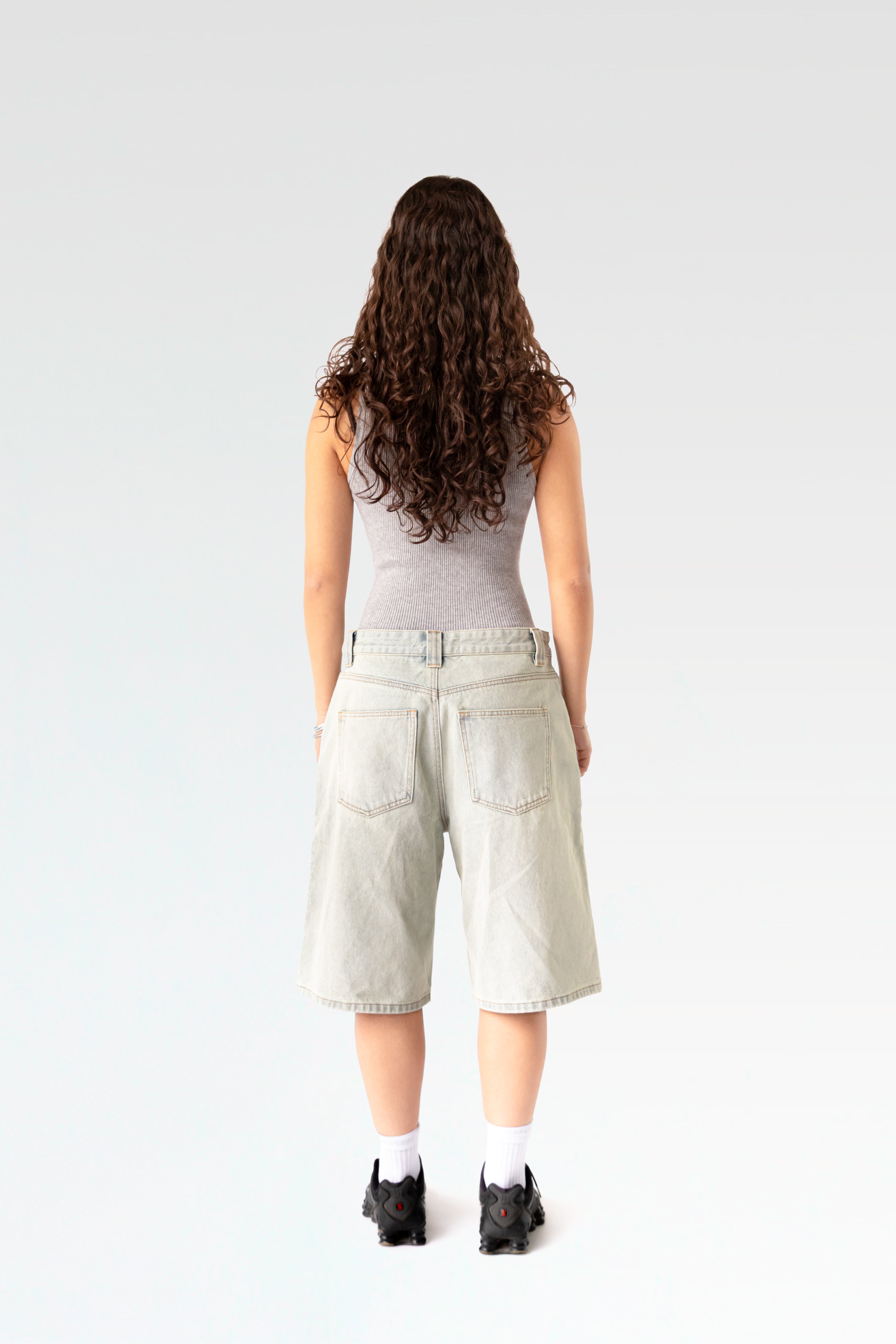 Rückansicht einer hellblauen, weit geschnittenen Jeansshorts aus gewaschenem Denim. Die Shorts reicht bis knapp übers Knie und zeigt zwei aufgesetzte Gesäßtaschen. Getragen von einer stehenden Person mit langen Locken, sportlichen Sneakers und weißem Tanktop.