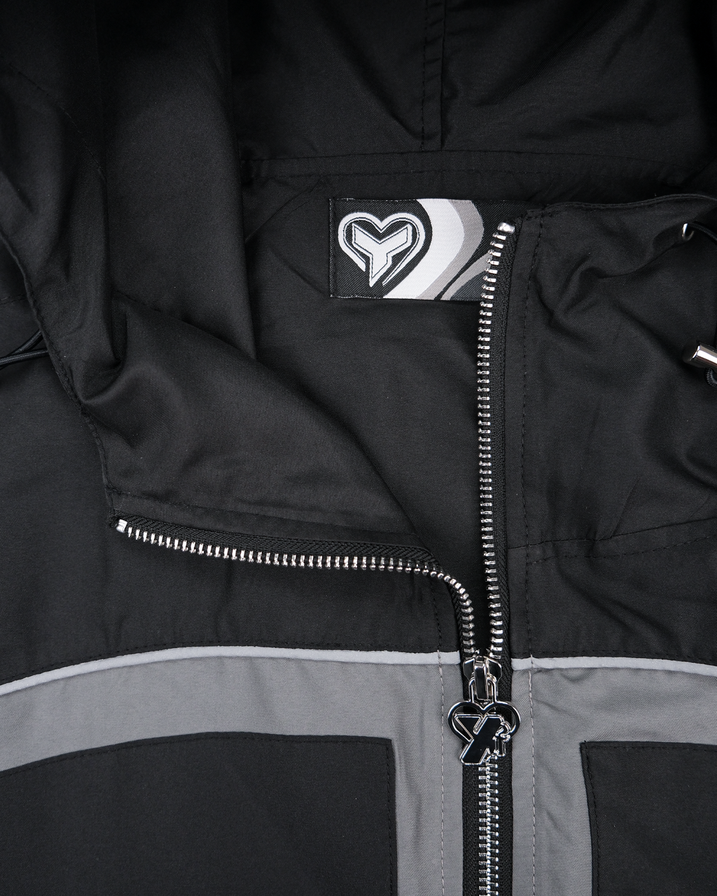 Detail der Innenseite der Jacke mit Label. Das Etikett zeigt ein stilisiertes Herzlogo mit grafischen Streifen. Der silberne Zipper trägt ebenfalls ein Herzsymbol mit integriertem „Y“-Buchstaben.

