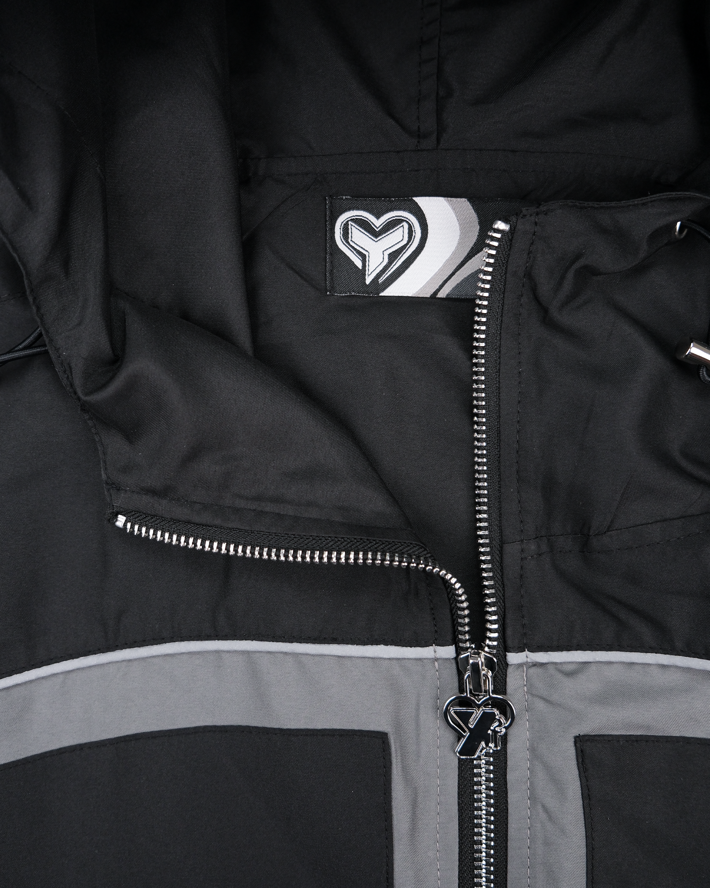 Detail der Innenseite der Jacke mit Label. Das Etikett zeigt ein stilisiertes Herzlogo mit grafischen Streifen. Der silberne Zipper trägt ebenfalls ein Herzsymbol mit integriertem „Y“-Buchstaben.

