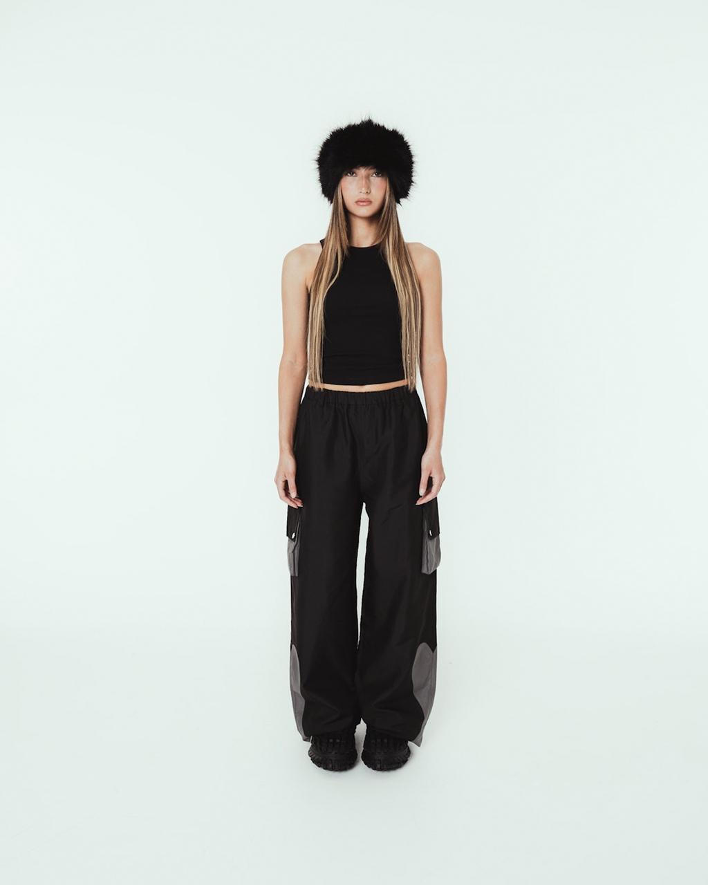 Person trägt eine schwarze, weit geschnittene Cargo-Hose mit elastischem Bund und grauen Kontrasteinsätzen an den Taschen und Beinabschlüssen. Die Hose hat seitliche Cargotaschen mit Druckknopfklappen sowie auffällige Einsätze im Kniebereich. Kombiniert mit schwarzem Croptop, Fellmütze und schwarzen Schuhen. Aufnahme in heller Studio-Umgebung.

