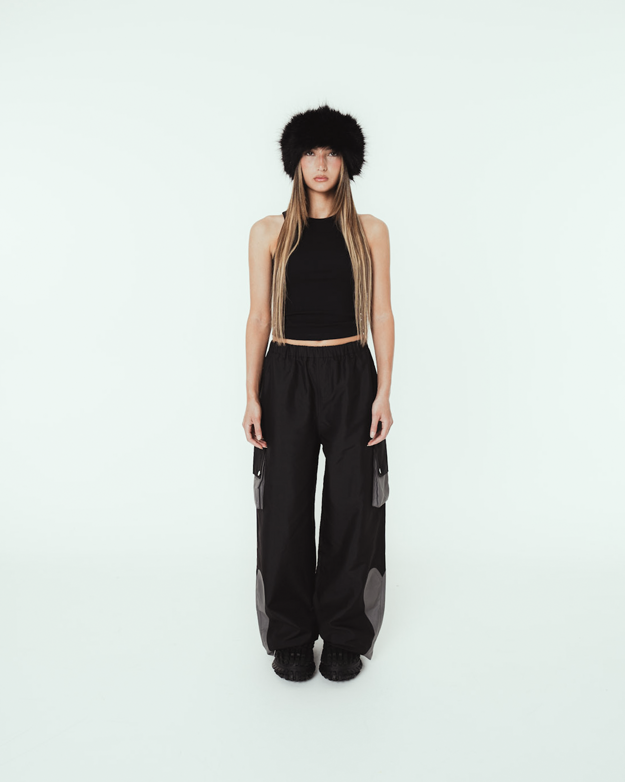 Person trägt eine schwarze, weit geschnittene Cargo-Hose mit elastischem Bund und grauen Kontrasteinsätzen an den Taschen und Beinabschlüssen. Die Hose hat seitliche Cargotaschen mit Druckknopfklappen sowie auffällige Einsätze im Kniebereich. Kombiniert mit schwarzem Croptop, Fellmütze und schwarzen Schuhen. Aufnahme in heller Studio-Umgebung.


