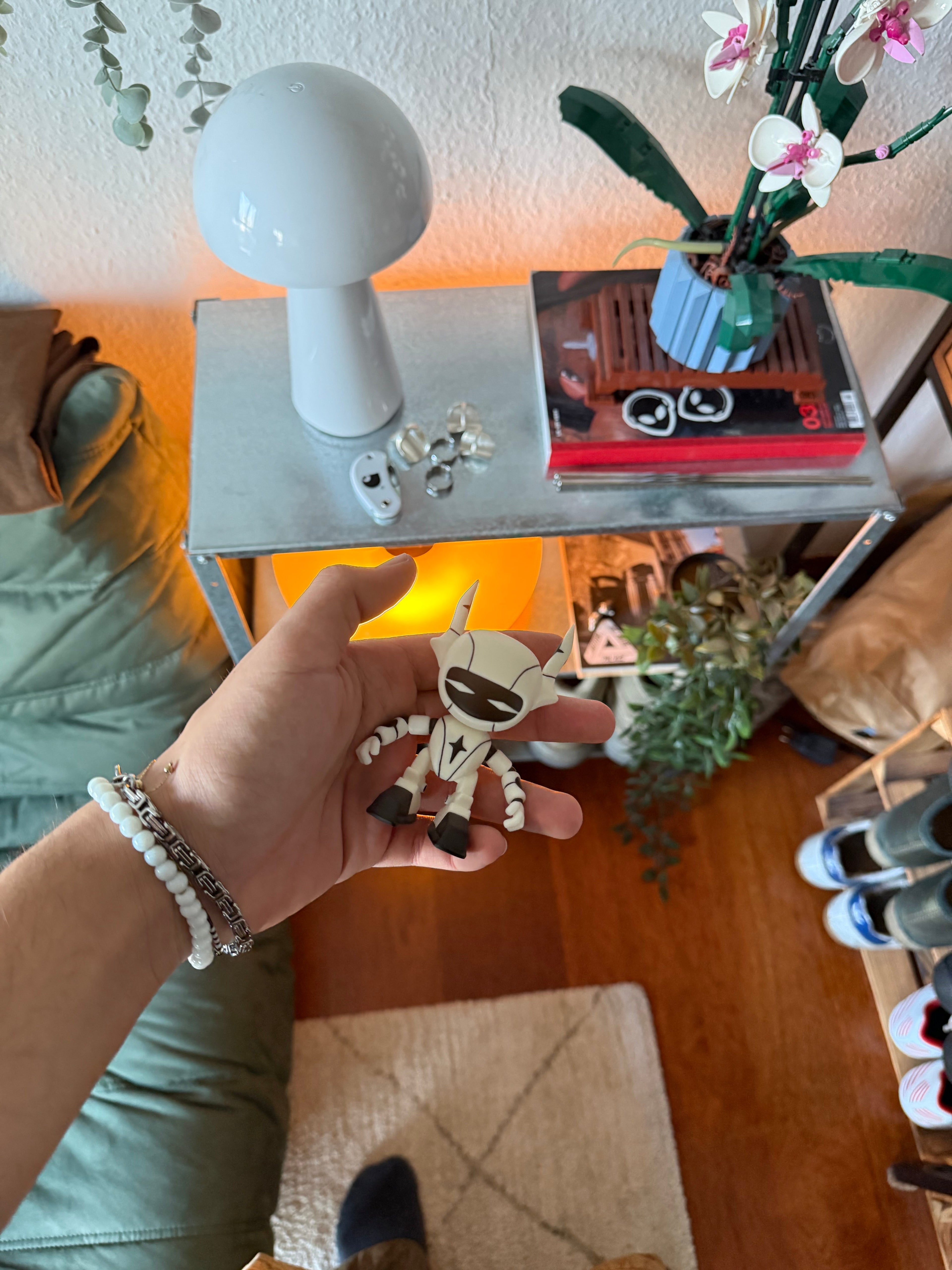 Eine Person hält die Roboterfigur in der linken Hand vor einem wohnlichen Hintergrund mit Tischlampe, Pflanzen und Regalen. Die Figur erscheint kompakt und handlich – etwa handgroß.