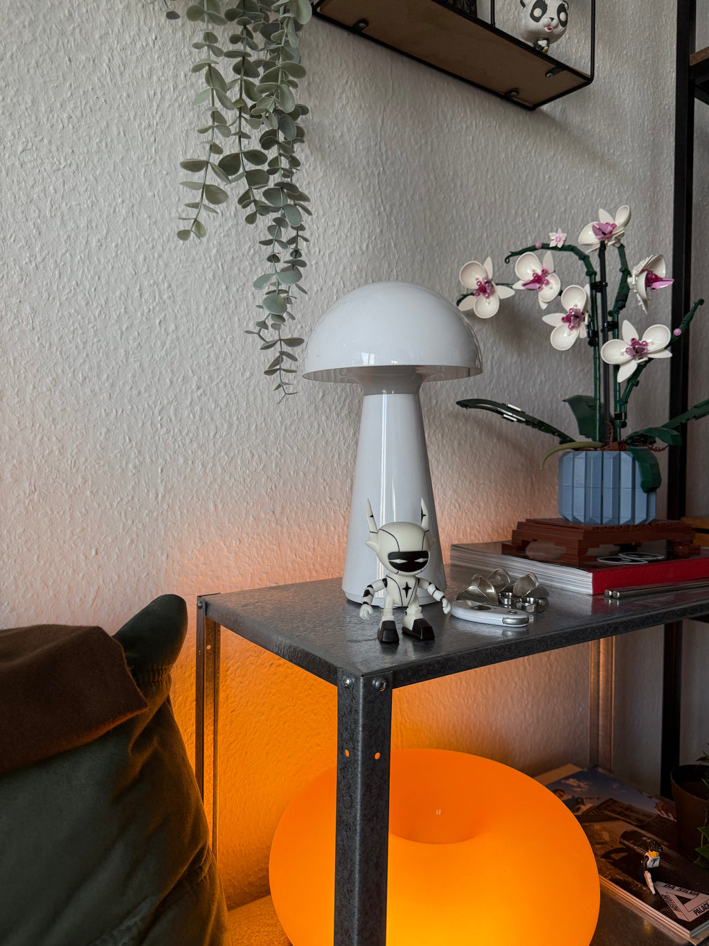 Die Figur steht dekorativ auf einem Metallregal neben einer weißen Pilzlampe, einem Schlüsselbund und einem Lego-Orchideen-Arrangement. Die Umgebung wirkt wohnlich und gemütlich, was den Einsatz als Dekoobjekt unterstreicht.