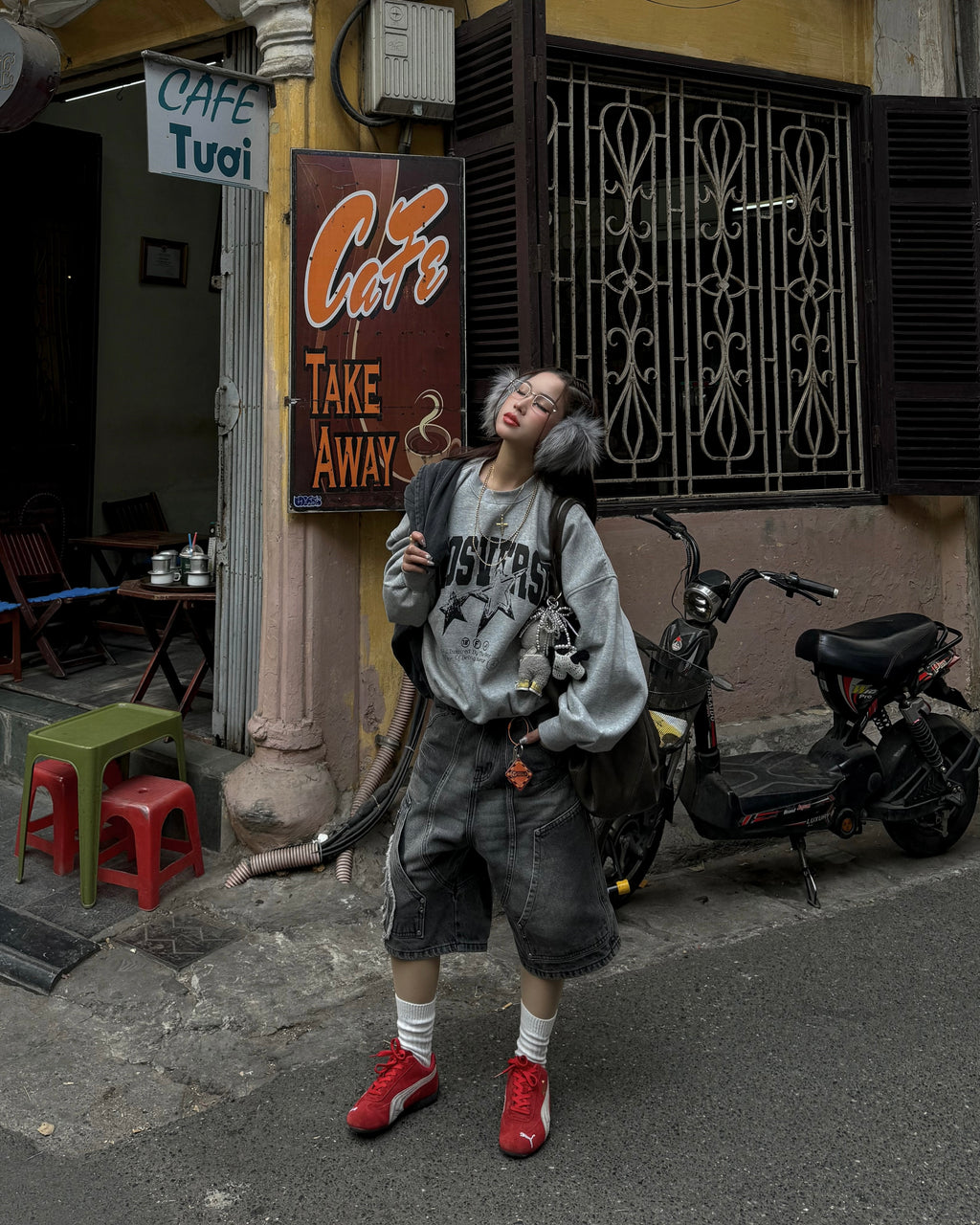 Straßenaufnahme einer Person mit grauer Oversize-Sweatjacke, Ohrenschützern und roten Turnschuhen. Die grauen Jeansshorts reichen bis knapp über die Knie und zeigen das auffällige Patch sowie die symmetrischen Frontnähte im Stil von Workwear.