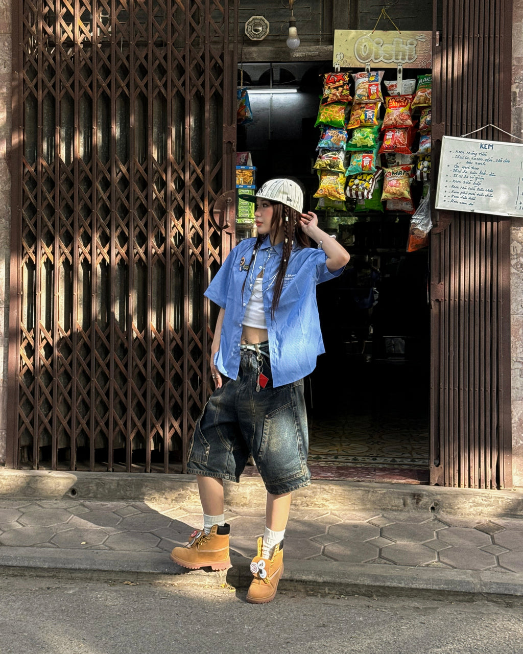Weit geschnittene Denim-Shorts mit ausgeprägter Waschung, getragen mit crop-top, offener Hemdbluse und Stiefeln. Die Shorts zeigen die auffällige Fransenapplikation und funktionale Taschen in urbanem Setting.