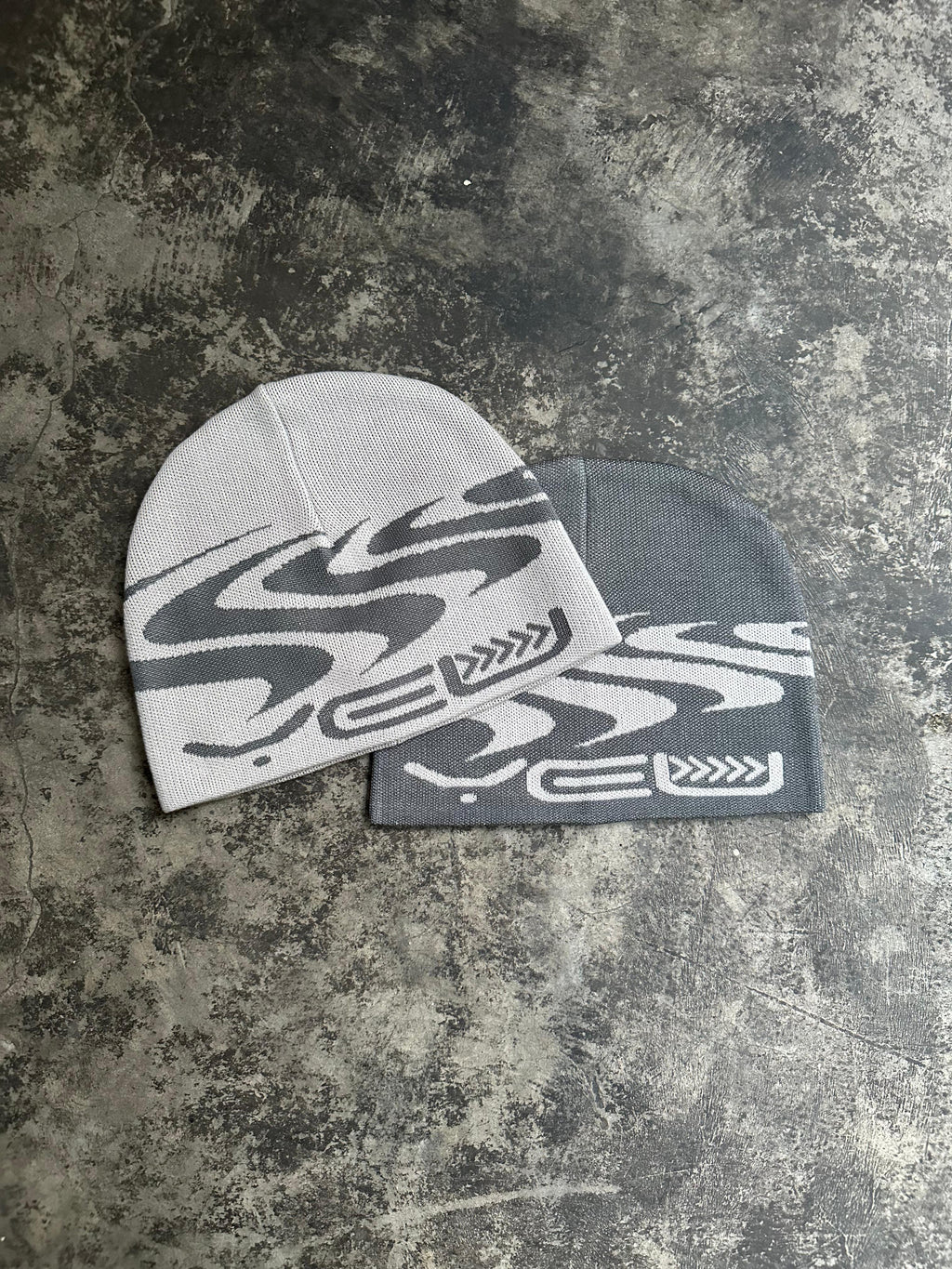  Zwei Beanies auf grauem Boden – oben eine beige Mütze mit grauem Wellenmotiv und „YEU“-Schriftzug, darunter eine dunkelgraue Variante mit gleichem Design.