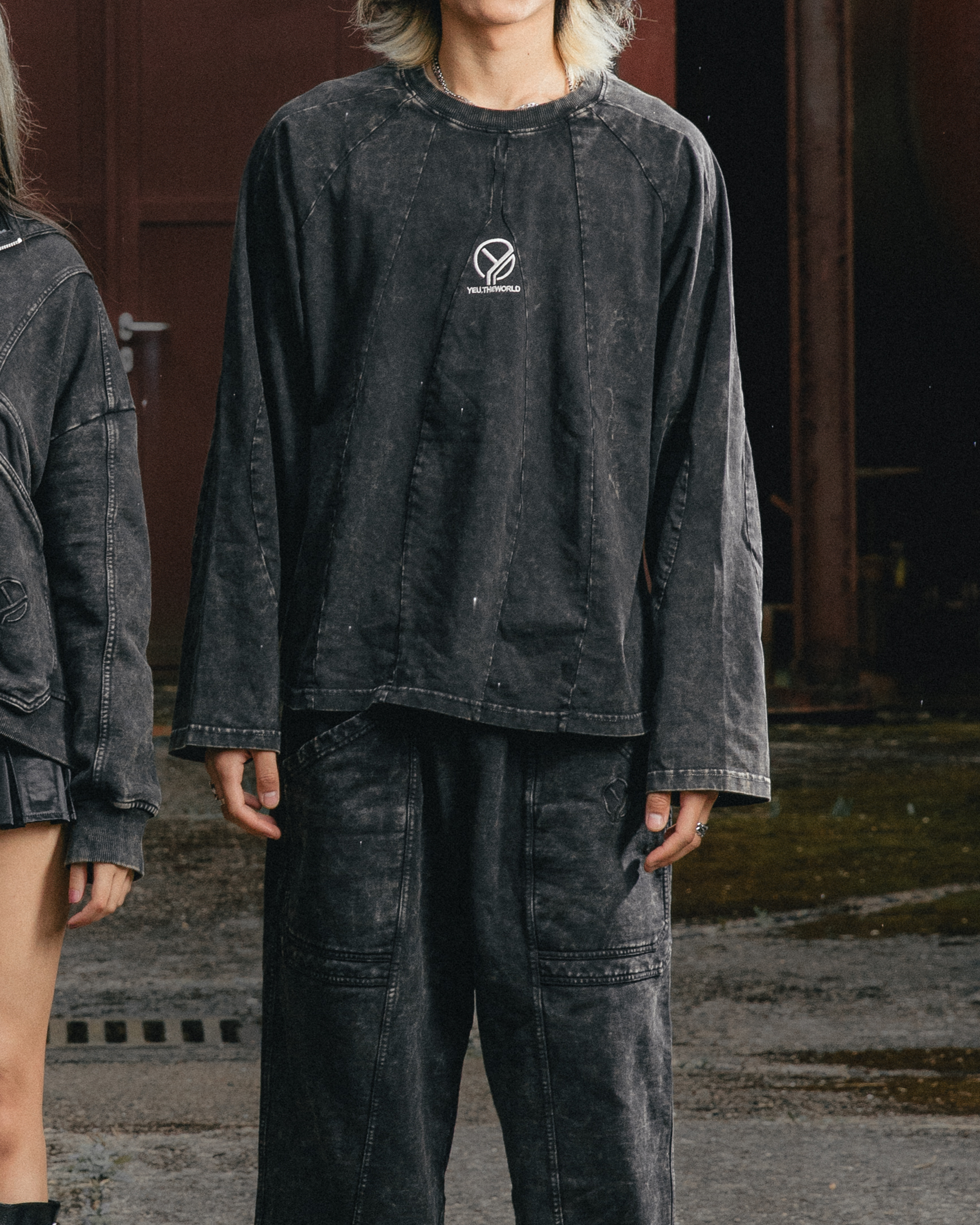 Tragebild in dunkler Urban-Umgebung: Eine Person trägt das langärmelige Shirt in verwaschenem Schwarz mit vertikalen Ziernähten und mittiger Logo-Stickerei „YEU.THEWORLD“. Das Shirt wirkt locker geschnitten und wird mit passender verwaschener Hose kombiniert.