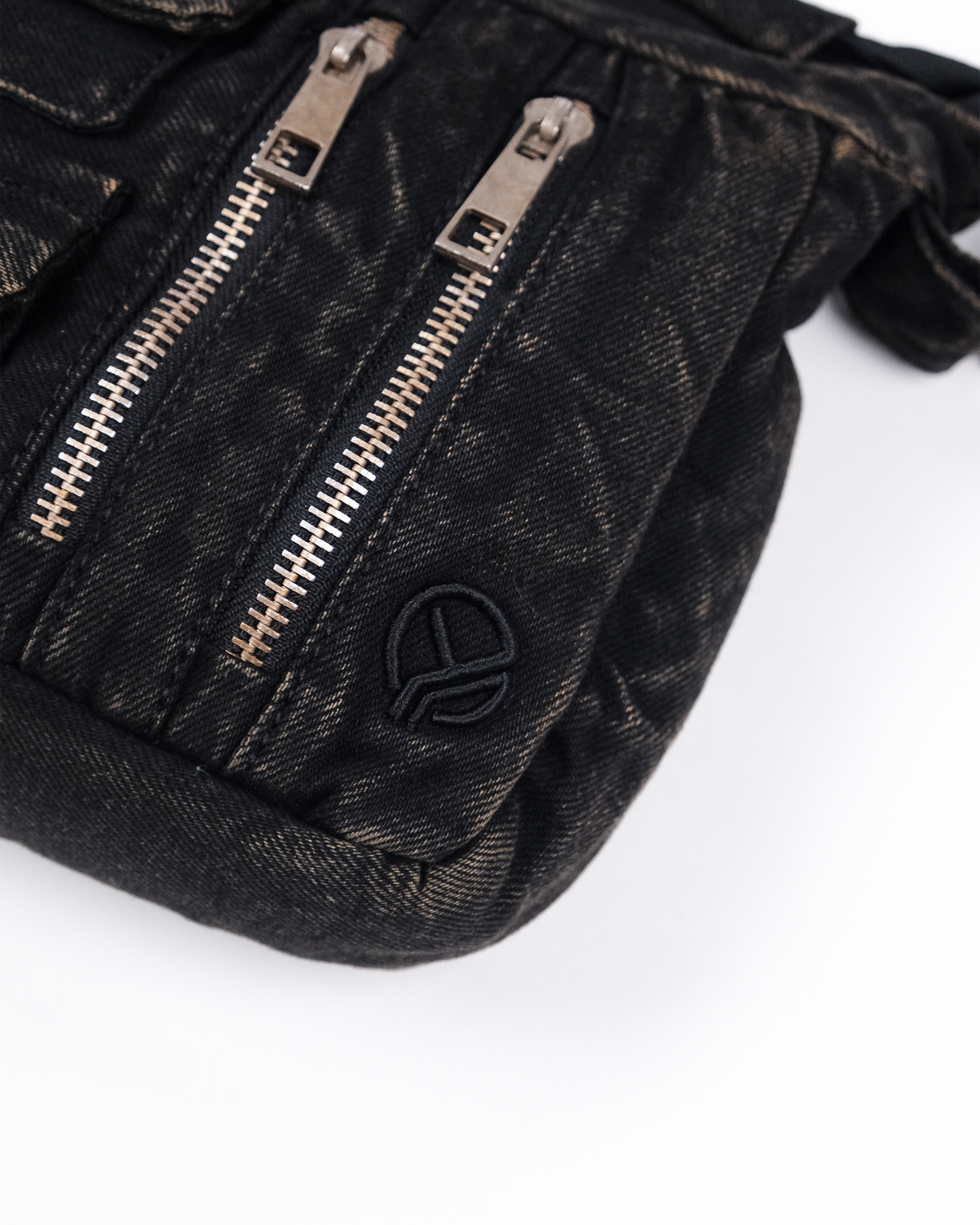 Nahaufnahme einer schwarzen Denim-Umhängetasche mit zwei silberfarbenen Reißverschlüssen und einem aufgestickten Logo in Schwarz in der rechten unteren Ecke.
