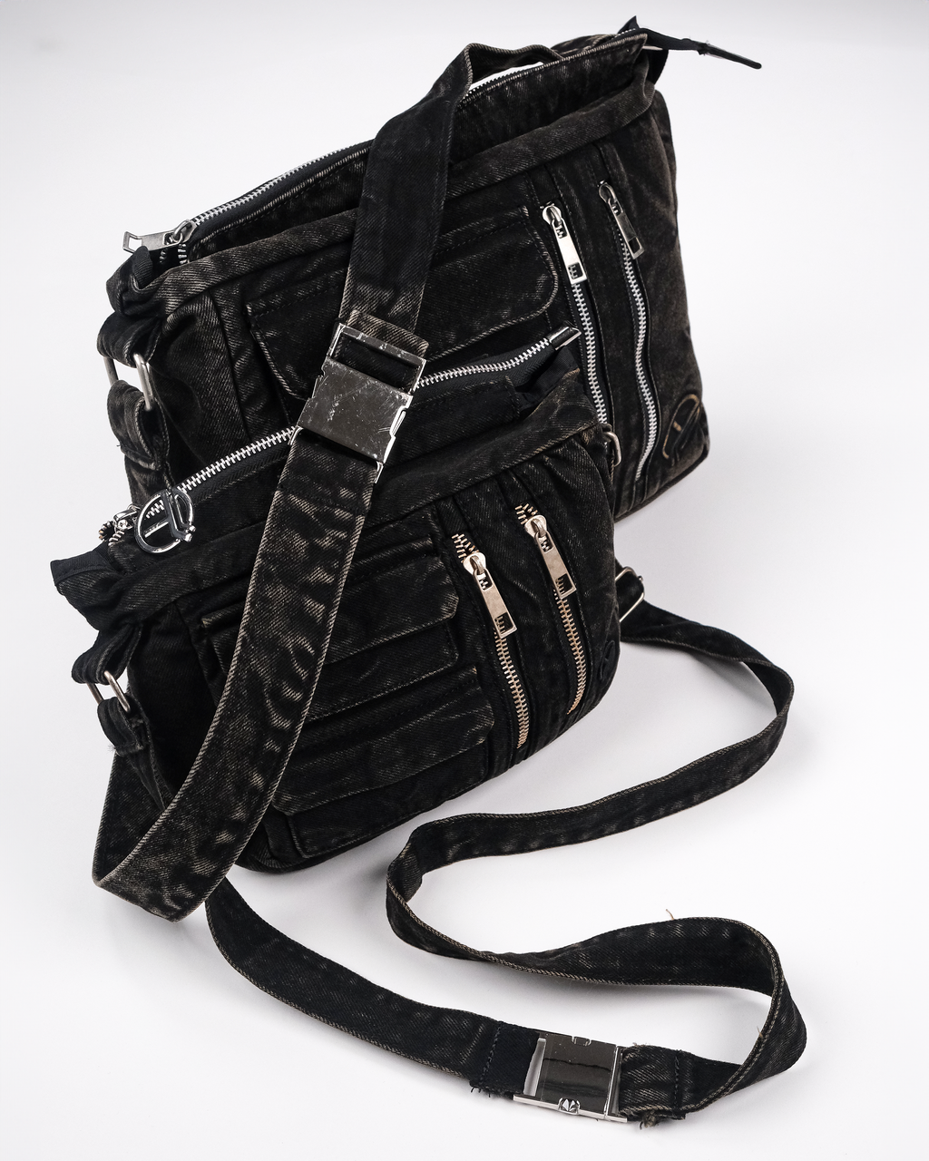 Zwei schwarze Umhängetaschen aus gewaschenem Denim, übereinandergelegt gezeigt. Sichtbar sind silberne Zipper, aufgesetzte Taschen und ein verstellbarer Schulterriemen mit Metallschnalle.
