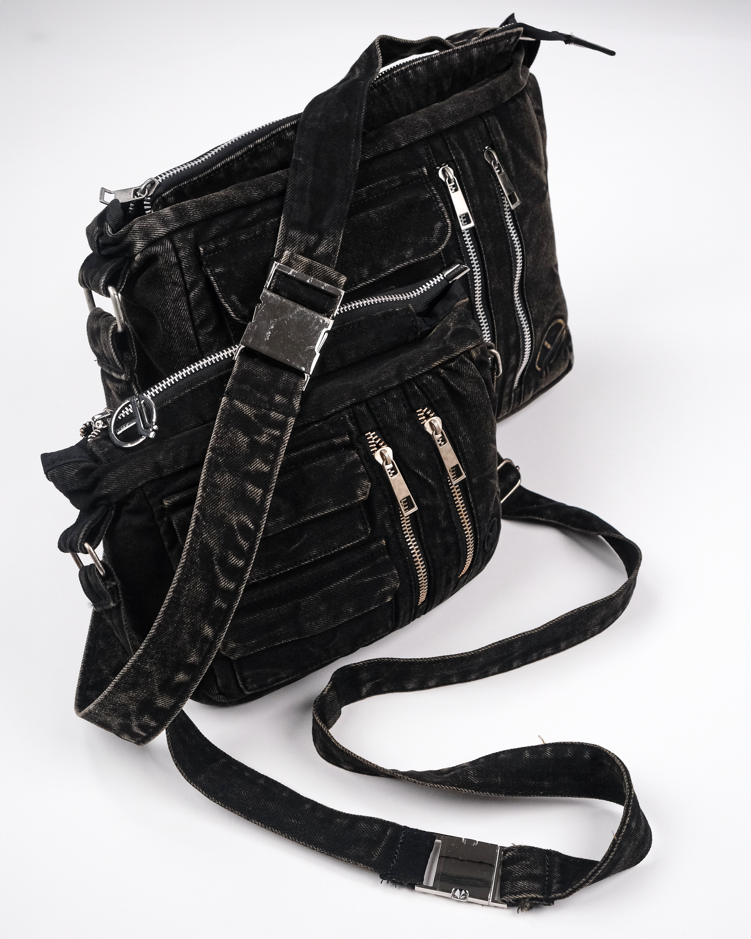 Zwei schwarze Umhängetaschen aus gewaschenem Denim, übereinandergelegt gezeigt. Sichtbar sind silberne Zipper, aufgesetzte Taschen und ein verstellbarer Schulterriemen mit Metallschnalle.