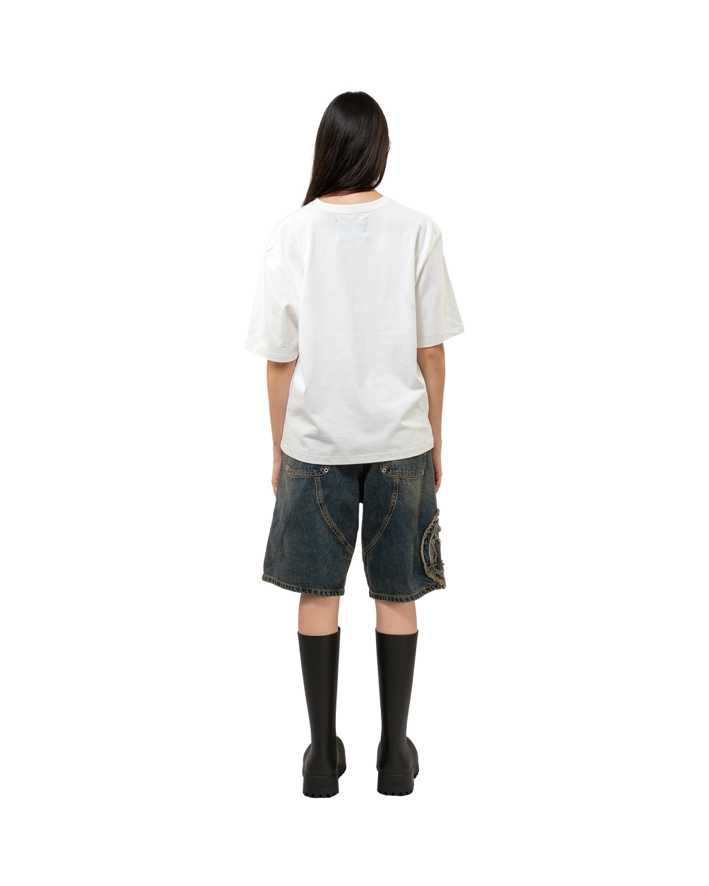 Denim-Shorts mit weiter, knielanger Passform, getragen von einer Person mit weißem Oversize-T-Shirt und kniehohen schwarzen Boots. Zu sehen ist die rückseitige Nahtführung und die Applikation am linken Hosenbein.