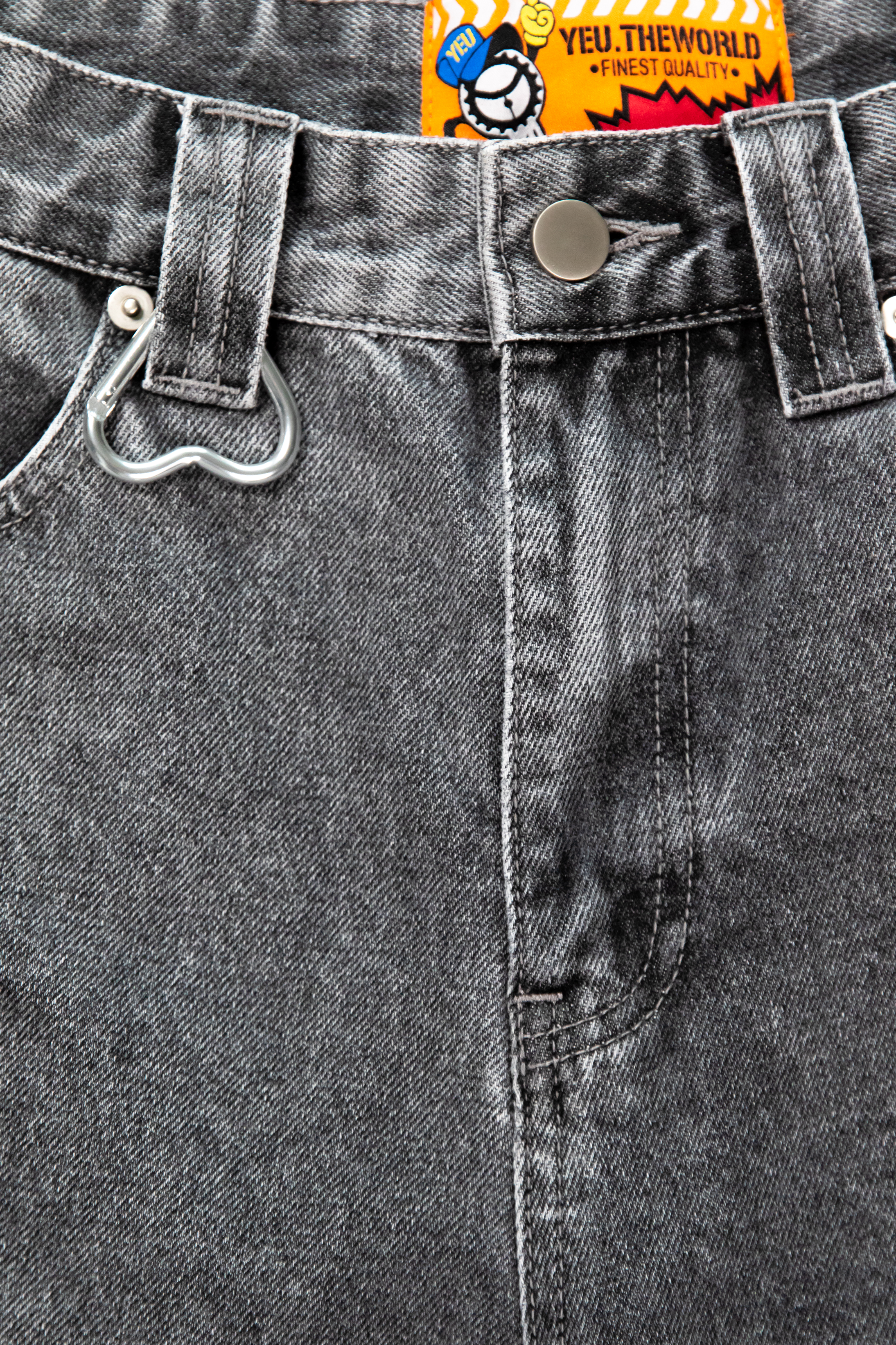 Detailaufnahme der Gürtelschlaufe mit silbernem Herz-Karabiner auf hellgrau verwaschenem Denim. Die grobe Stoffstruktur und Nahtverarbeitung sind sichtbar.