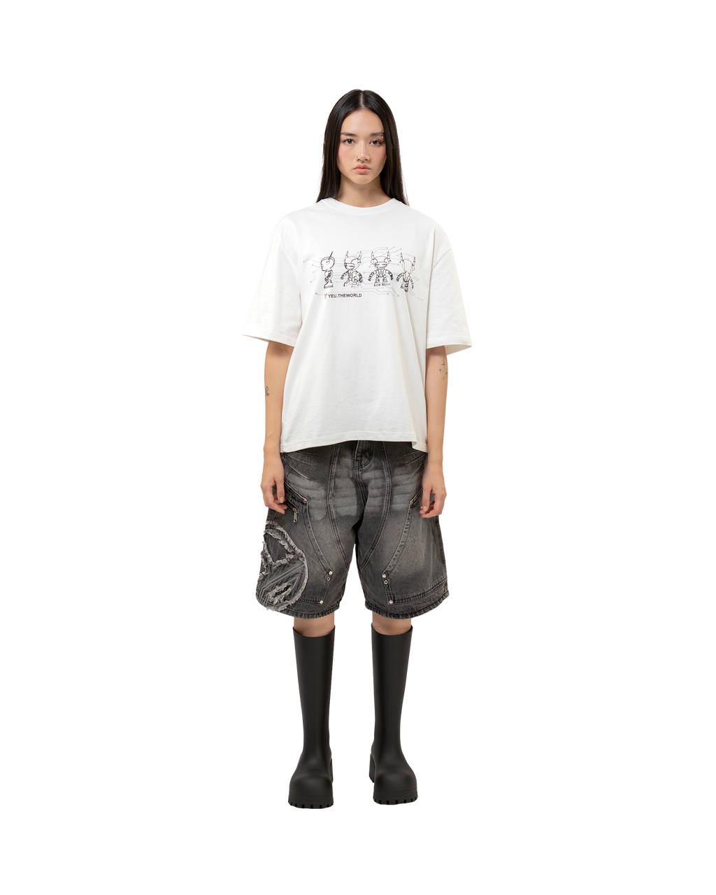 Frontansicht derselben Person in neutraler Pose mit weißem T-Shirt und knielangen grauen Jeansshorts. Sichtbar sind das große, ausgefranste Patch mit hellen Stickereien sowie der schräge Zipper auf dem linken Oberschenkel.
