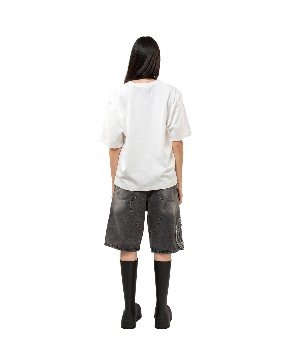 Rückansicht einer stehenden Person mit langen schwarzen Haaren in schwarzer Gummistiefel-Optik. Die grauen Jeansshorts enden unterhalb der Knie und zeigen auf der Rückseite die charakteristische Nahtführung sowie das sichtbare grafische Patch.