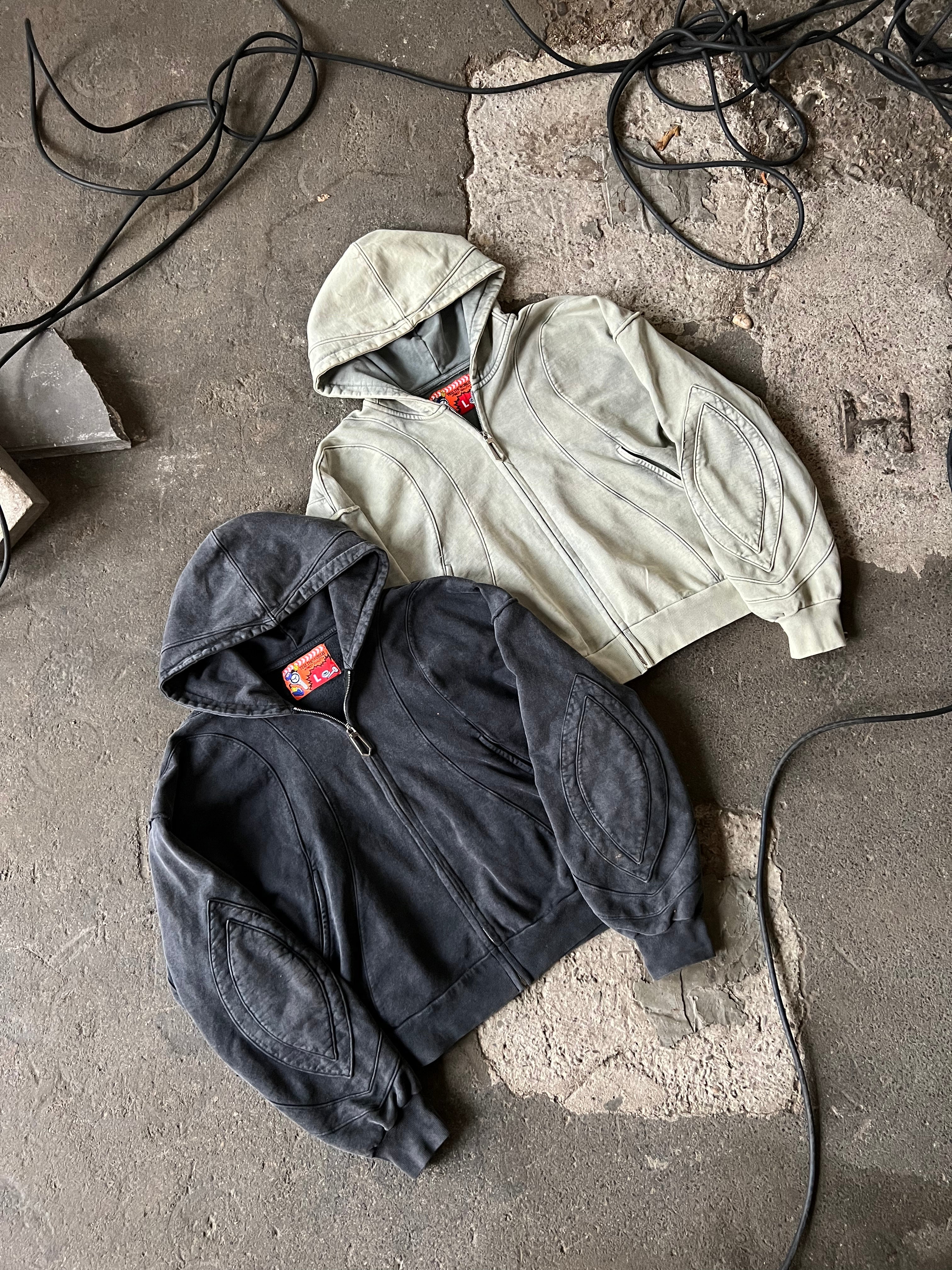 Der schwarze Zip-Hoodie liegt neben dem gleichen Modell in hellem Grün auf grauem Untergrund. Beide zeigen markante Liniennähte auf Brust und Ärmeln sowie die roten Nackenlabels mit Grafikmotiv. Die Struktur und Waschung des Stoffs sind gut erkennbar.