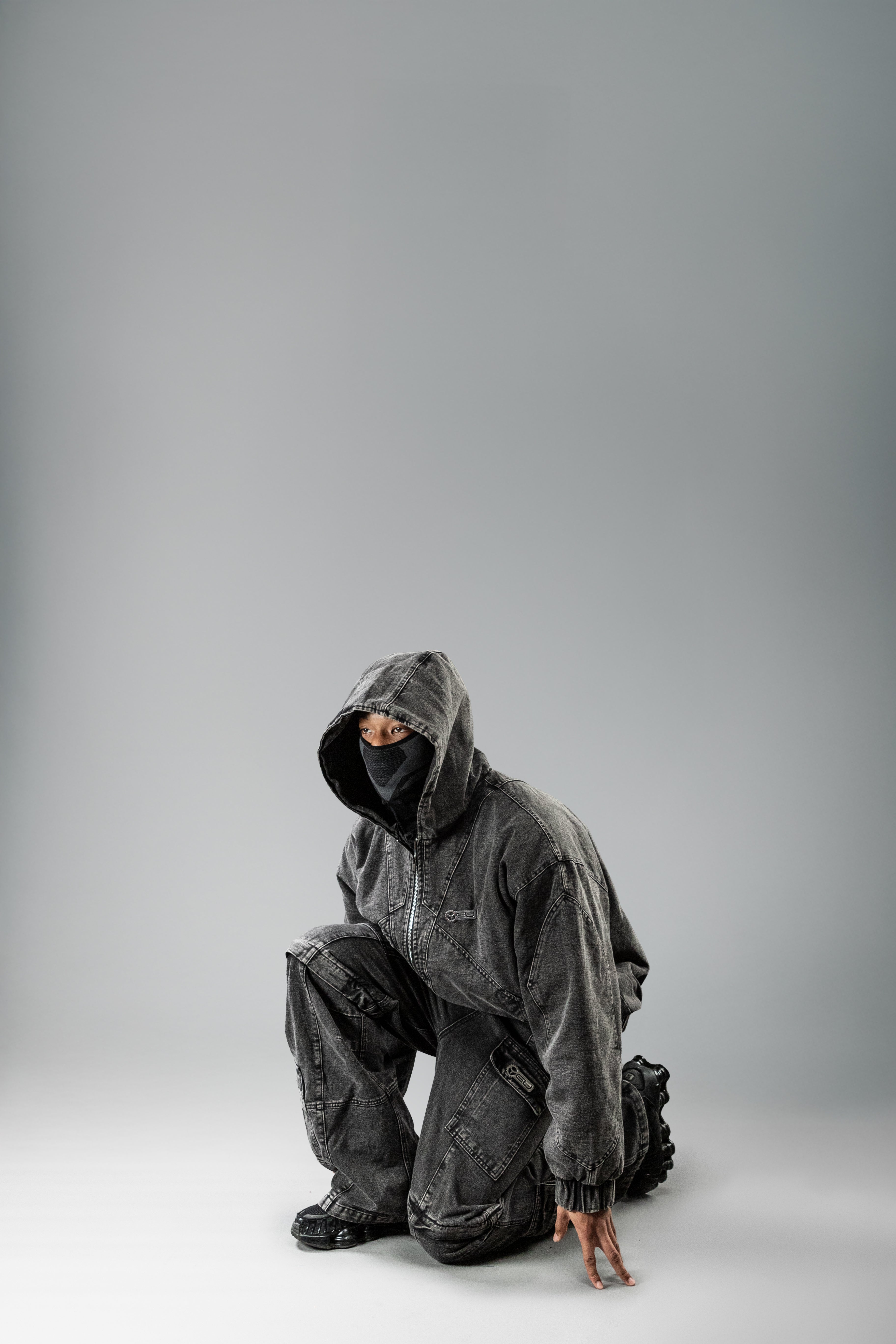 Person in dunkler Denim-Kapuzenjacke und passender Hose in geduckter Pose. Die Kapuze ist über den Kopf gezogen, das Gesicht ist teilweise durch eine schwarze Gesichtsmaske verdeckt.