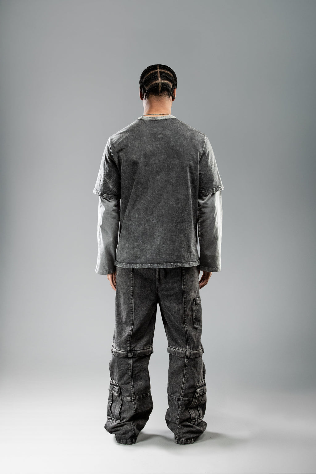 Rückansicht des schwarzen Layering-Shirts im getragenen Zustand. Das Shirt hat einen geraden Saum und hellgraue Langarmärmel, die unter dem kurzen T-Shirt-Oberteil hervorstehen. Getragen mit verwaschener schwarzer Cargohose.