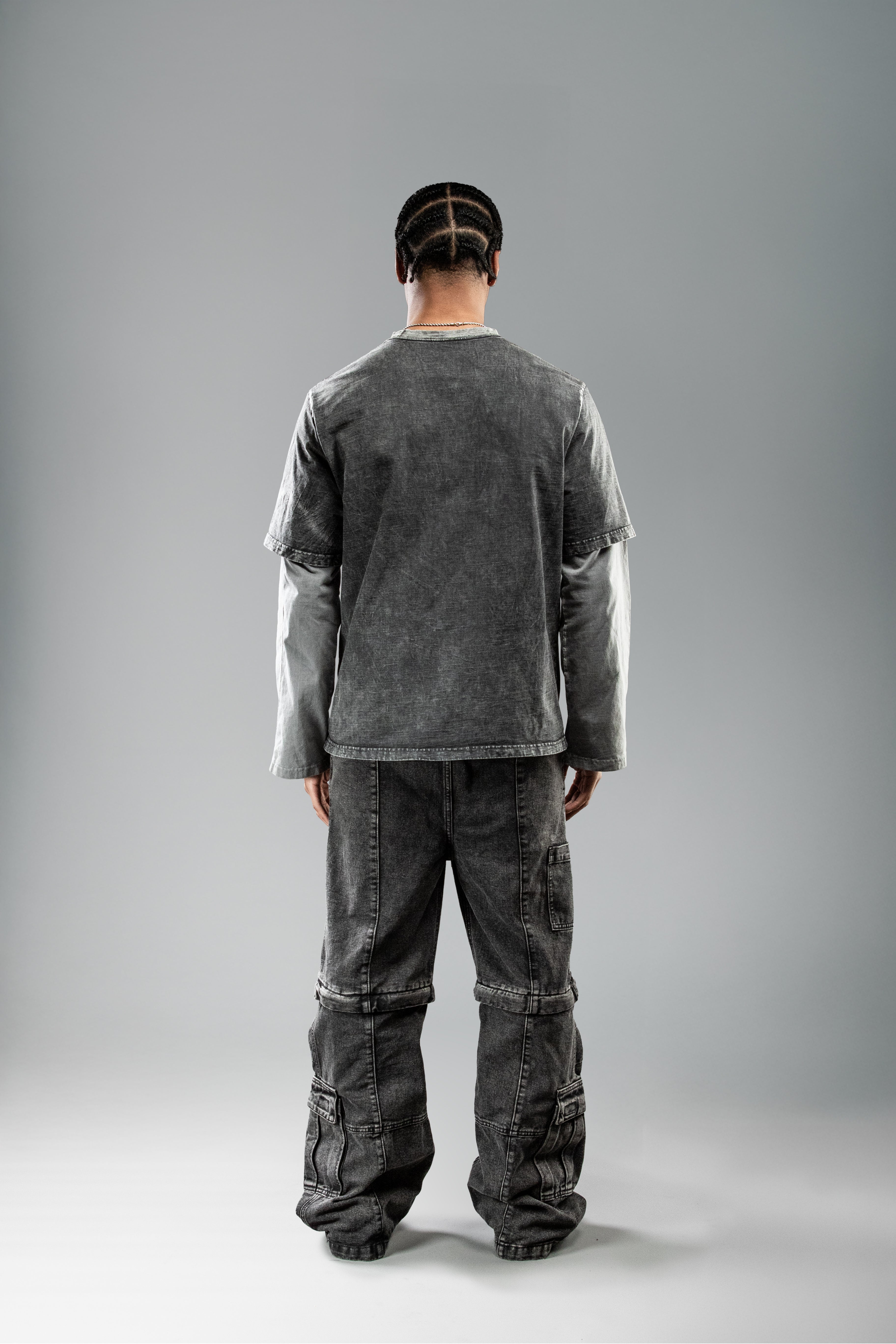 Rückansicht des schwarzen Layering-Shirts im getragenen Zustand. Das Shirt hat einen geraden Saum und hellgraue Langarmärmel, die unter dem kurzen T-Shirt-Oberteil hervorstehen. Getragen mit verwaschener schwarzer Cargohose.