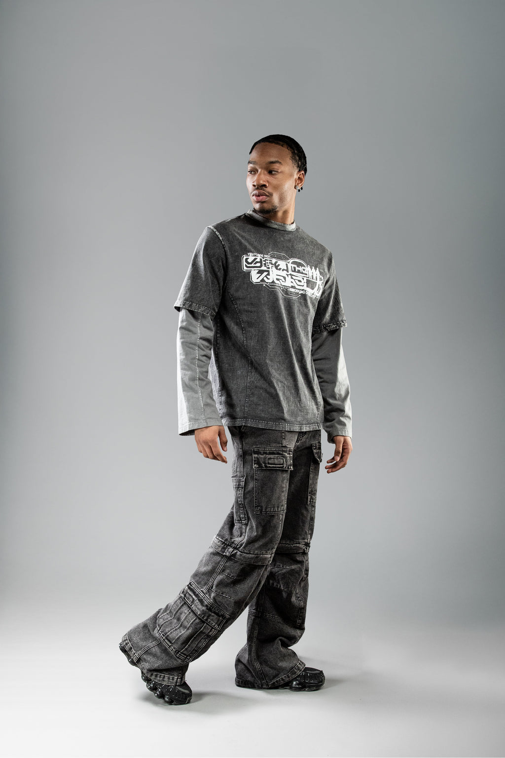 Dynamische Trageaufnahme des schwarzen Layering-Shirts. Die Person geht leicht gedreht in Bewegung, wodurch das Shirt, die grauen Ärmel und die lockere Passform der Cargohose gut sichtbar sind.