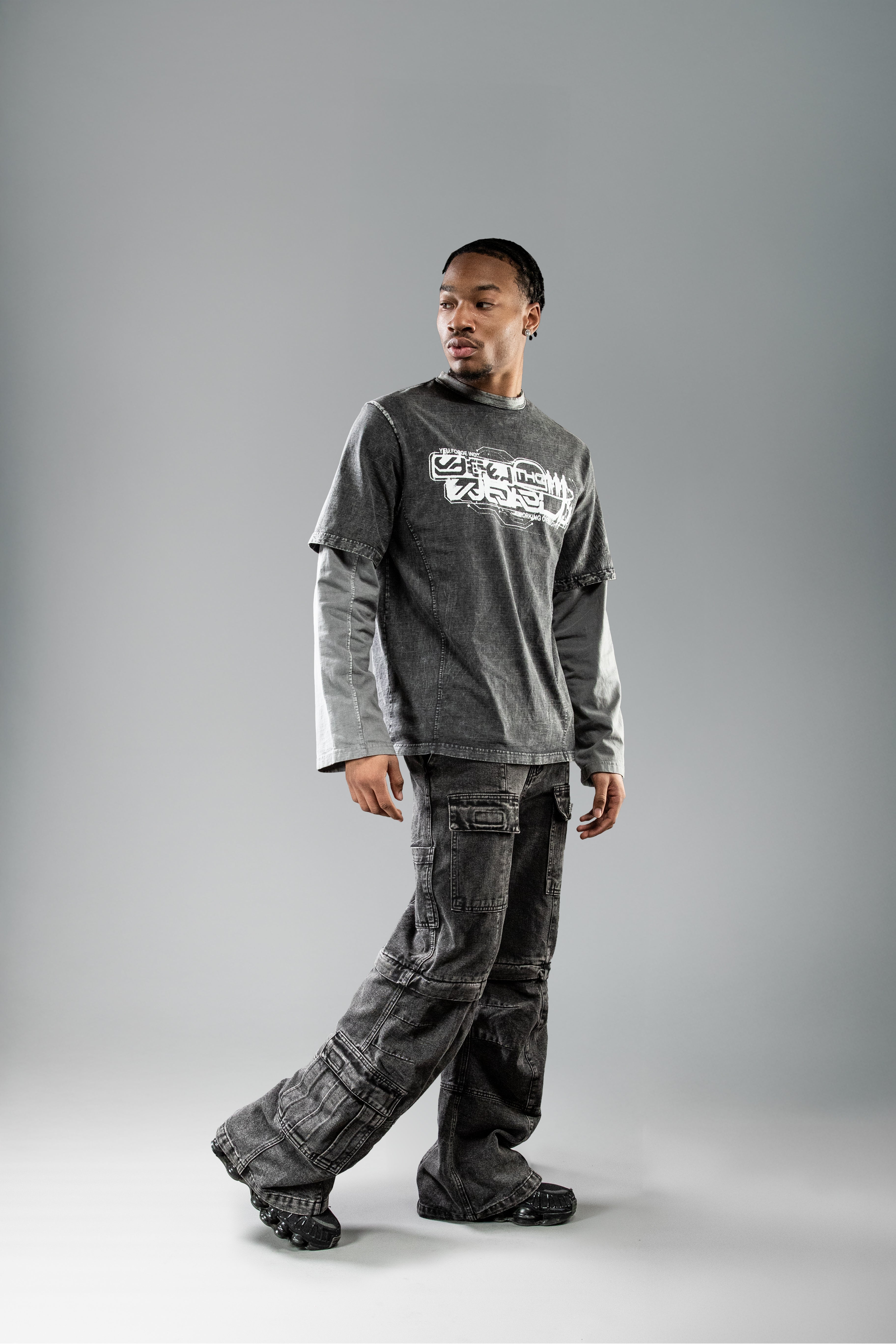 Dynamische Trageaufnahme des schwarzen Layering-Shirts. Die Person geht leicht gedreht in Bewegung, wodurch das Shirt, die grauen Ärmel und die lockere Passform der Cargohose gut sichtbar sind.