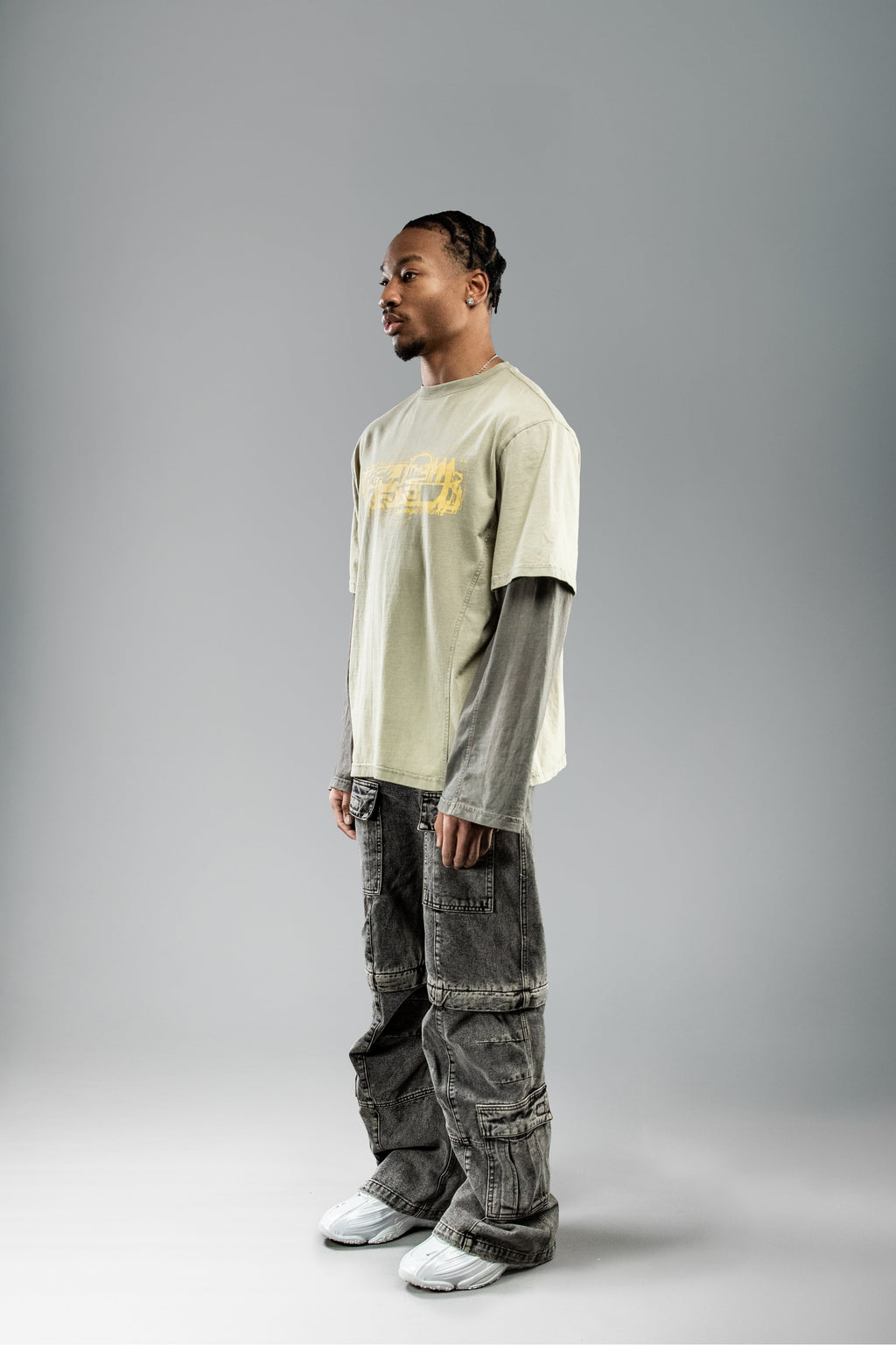 Seitlich-frontal aufgenommene Ansicht eines Models im olivgrünen Layer-T-Shirt mit gelbem Frontprint und integrierten langen Ärmeln in dunklem Grau. Kombiniert mit weiter, grauer Cargohose.