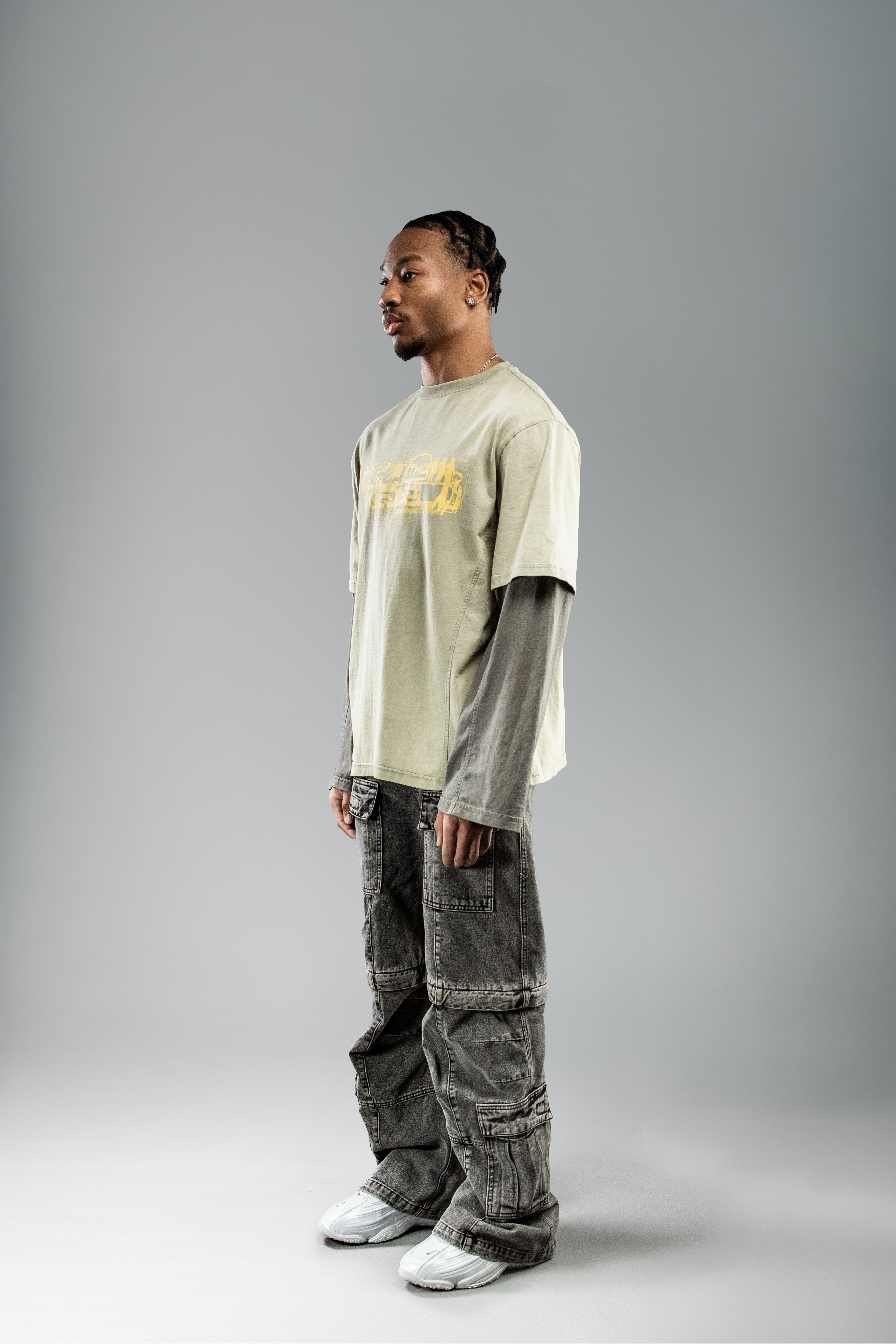 Seitlich-frontal aufgenommene Ansicht eines Models im olivgrünen Layer-T-Shirt mit gelbem Frontprint und integrierten langen Ärmeln in dunklem Grau. Kombiniert mit weiter, grauer Cargohose.