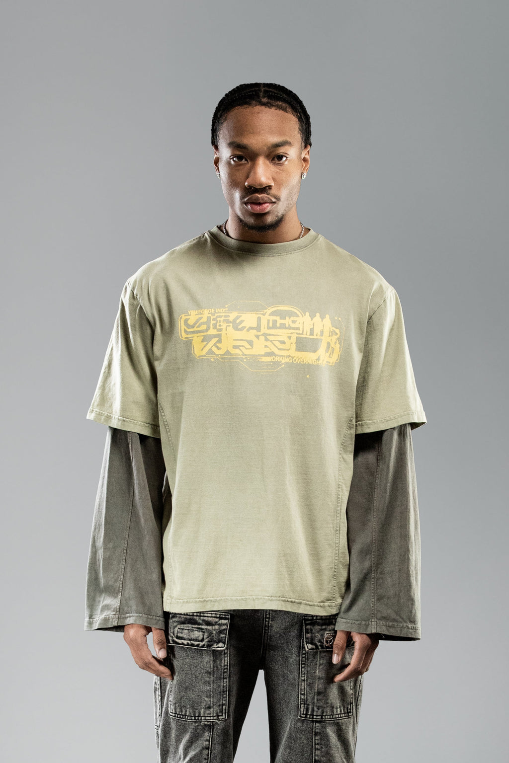 Frontale Nahaufnahme eines stehenden Models mit olivgrünem Layer-T-Shirt. Der gelbe, technisch wirkende Schriftzug auf der Brust ist zentriert platziert, darunter steht klein „WORKING OVERNIGHT“.