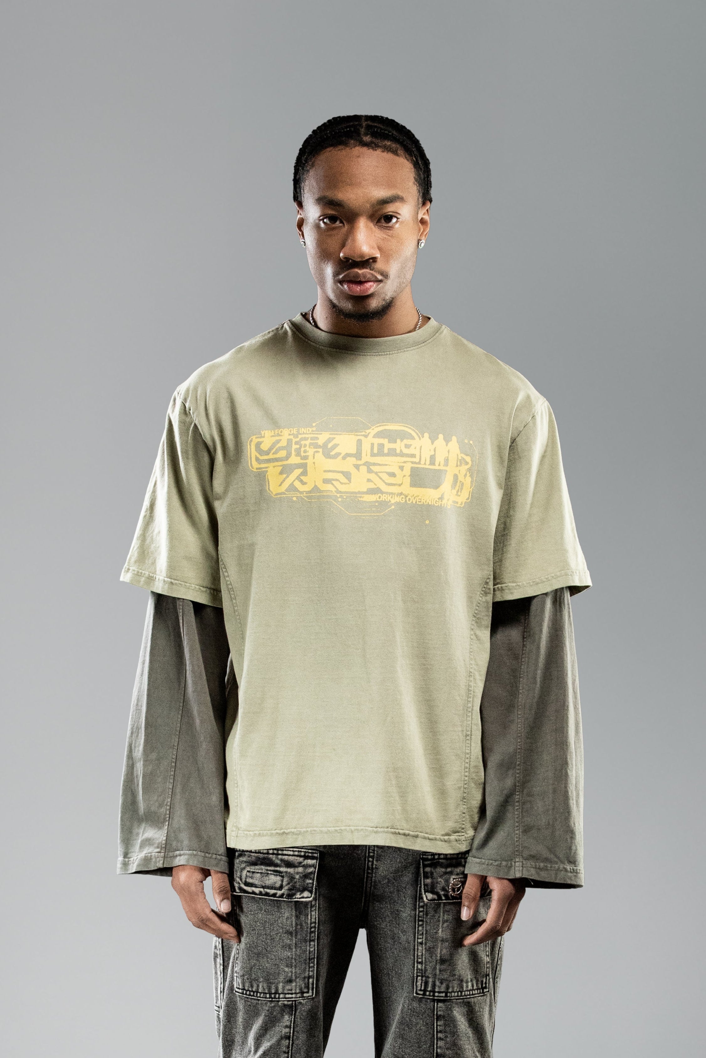 Frontale Nahaufnahme eines stehenden Models mit olivgrünem Layer-T-Shirt. Der gelbe, technisch wirkende Schriftzug auf der Brust ist zentriert platziert, darunter steht klein „WORKING OVERNIGHT“.
