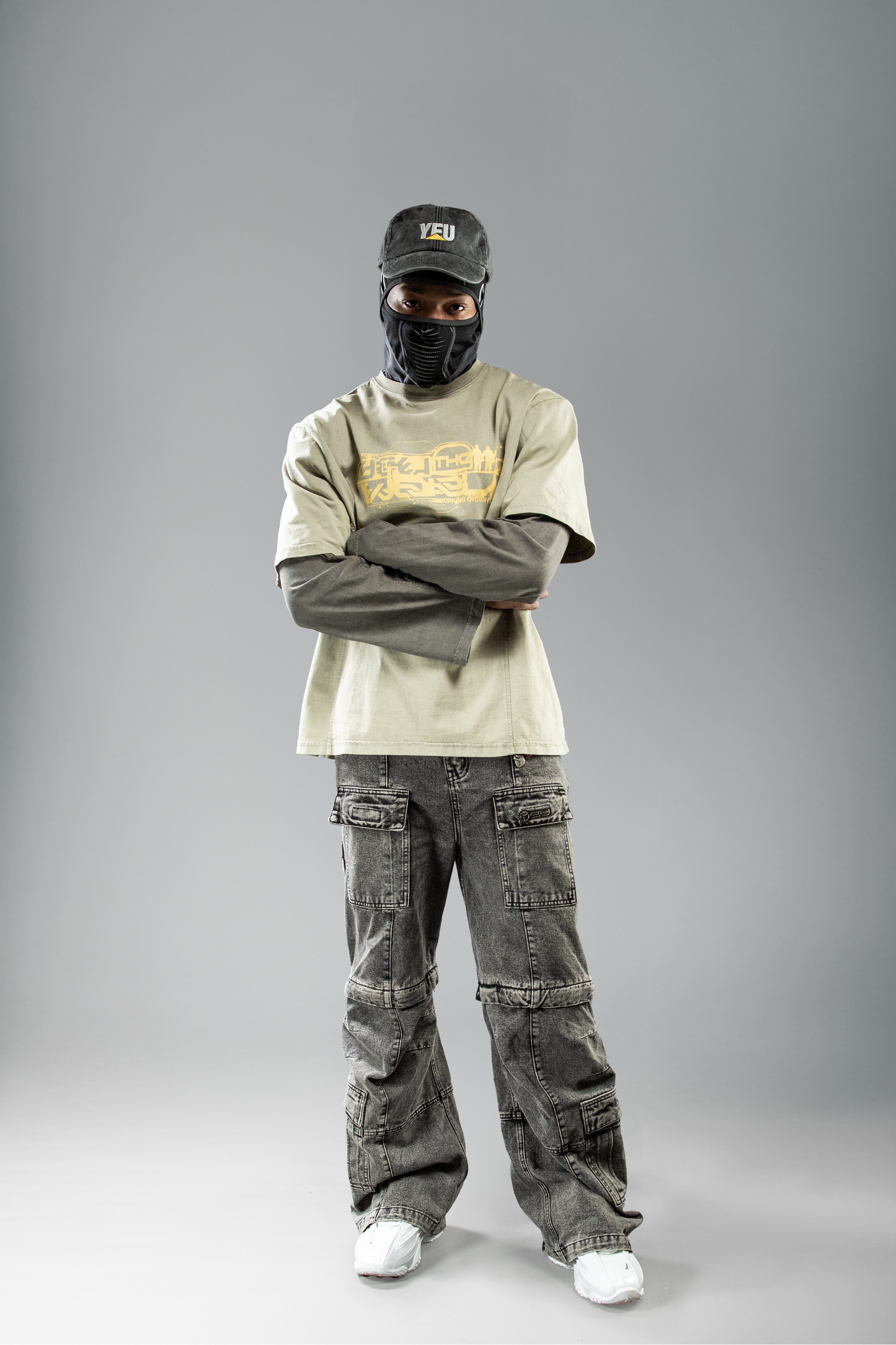 Frontale Ganzkörperaufnahme eines Models im olivgrünen Layer-Shirt mit überkreuzten Armen. Ergänzt durch schwarze Kappe mit „YEU“-Schriftzug, dunkle Gesichtsmaske und weite Cargohose.