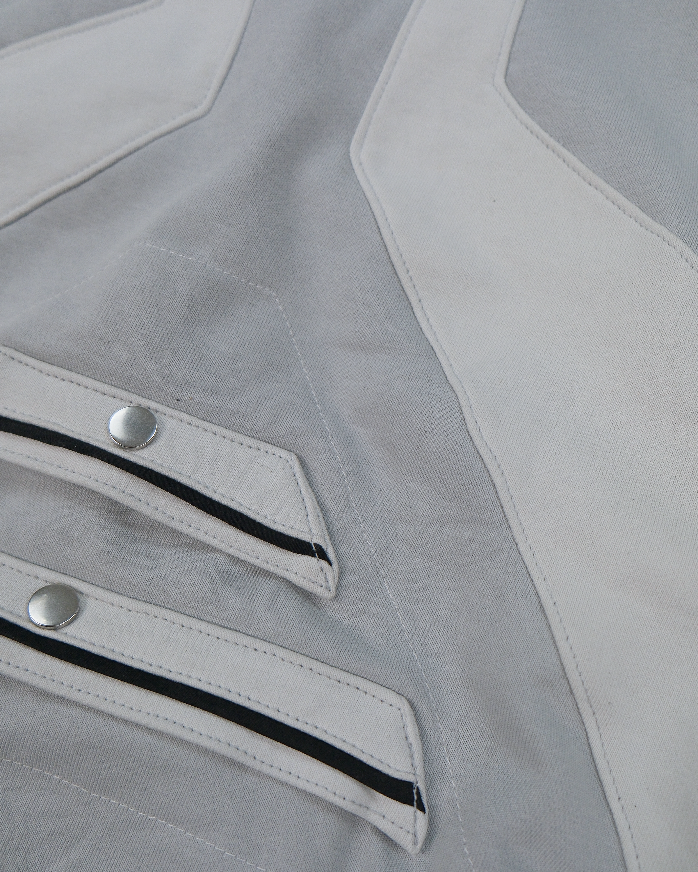 Hellgrauer Zip-Hoodie mit Kapuze, symmetrischem Einsatz in helleren Grautönen und auffälligem Reißverschluss in der Mitte. Die geometrisch angeordneten Stoffbahnen auf der Vorderseite formen ein auffächerndes Design.