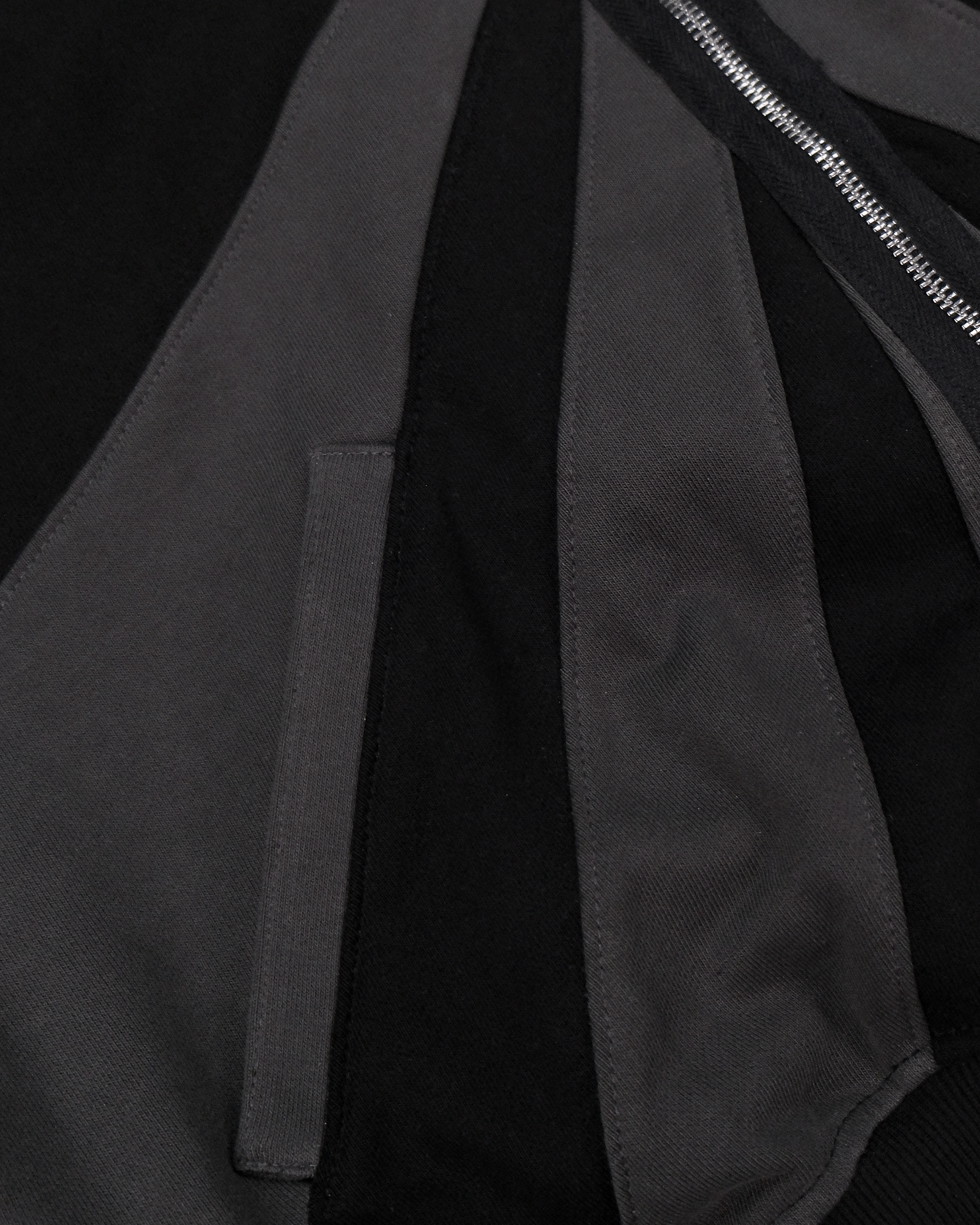 Vorderansicht einer schwarzen Kapuzenjacke mit geometrischer Panel-Konstruktion in dunklem Grau. Die Jacke besitzt einen durchgehenden Reißverschluss mit silbernem Stern-Zipper und breite Einsätze entlang des Rumpfes und der Ärmel.