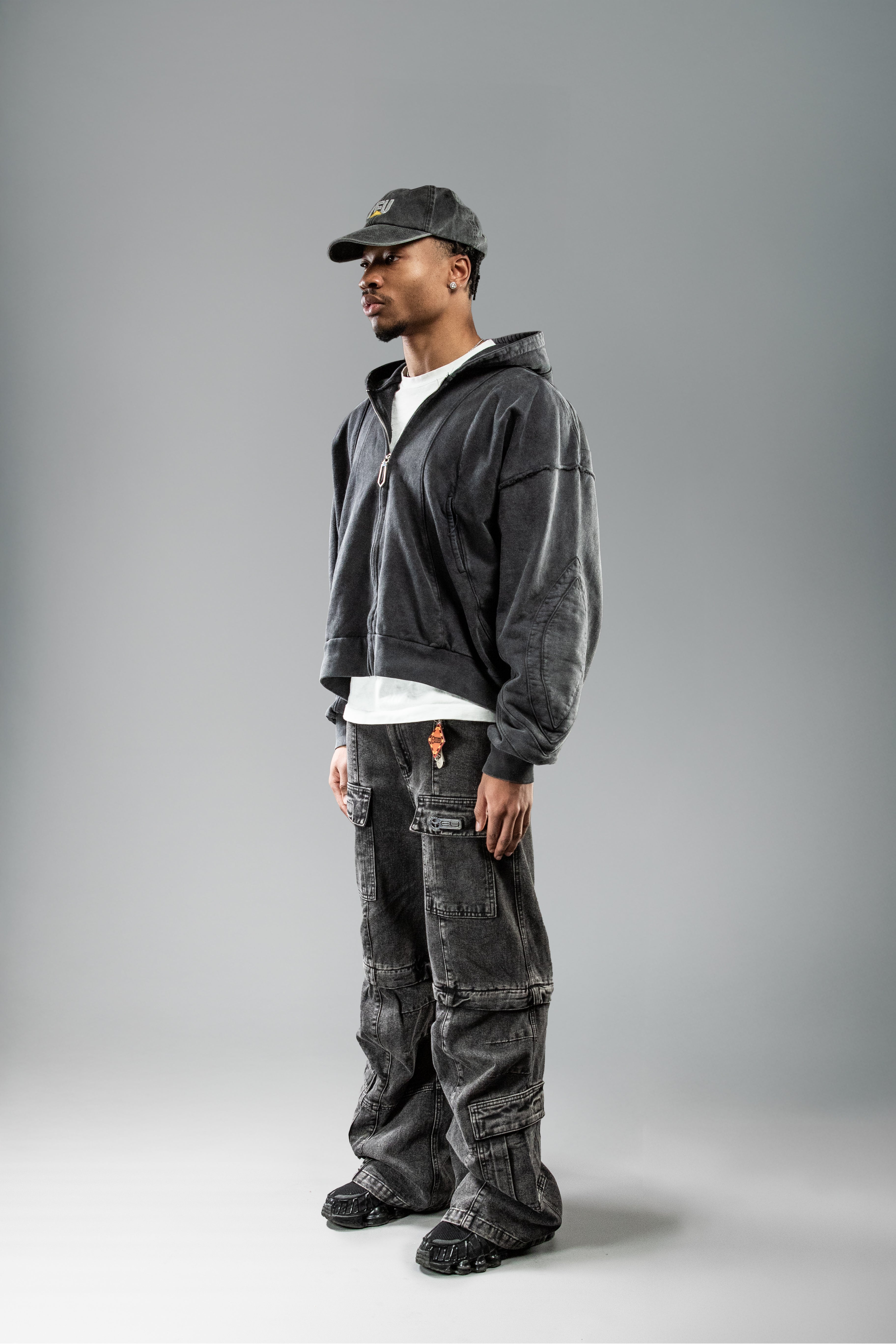 Schwarzer Zip-Hoodie leicht seitlich getragen. Die geformten Teilungsnähte auf Brust und Schulter heben sich durch die Waschung deutlich ab. Die Kapuze liegt nach hinten gefaltet.