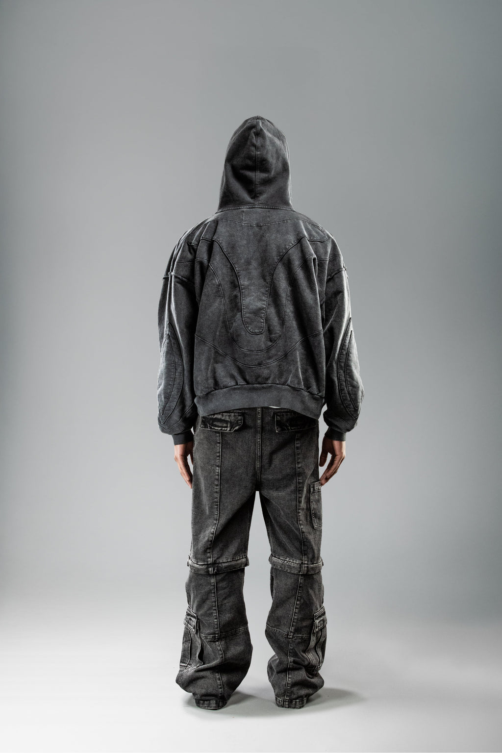 Getragene Rückenansicht des schwarzen Hoodies mit aufgesetzter Kapuze. Die Rückseite zeigt das geschwungene Nahtmuster in betonter Linienführung. Der Stoff wirkt leicht strukturiert und verwaschen.