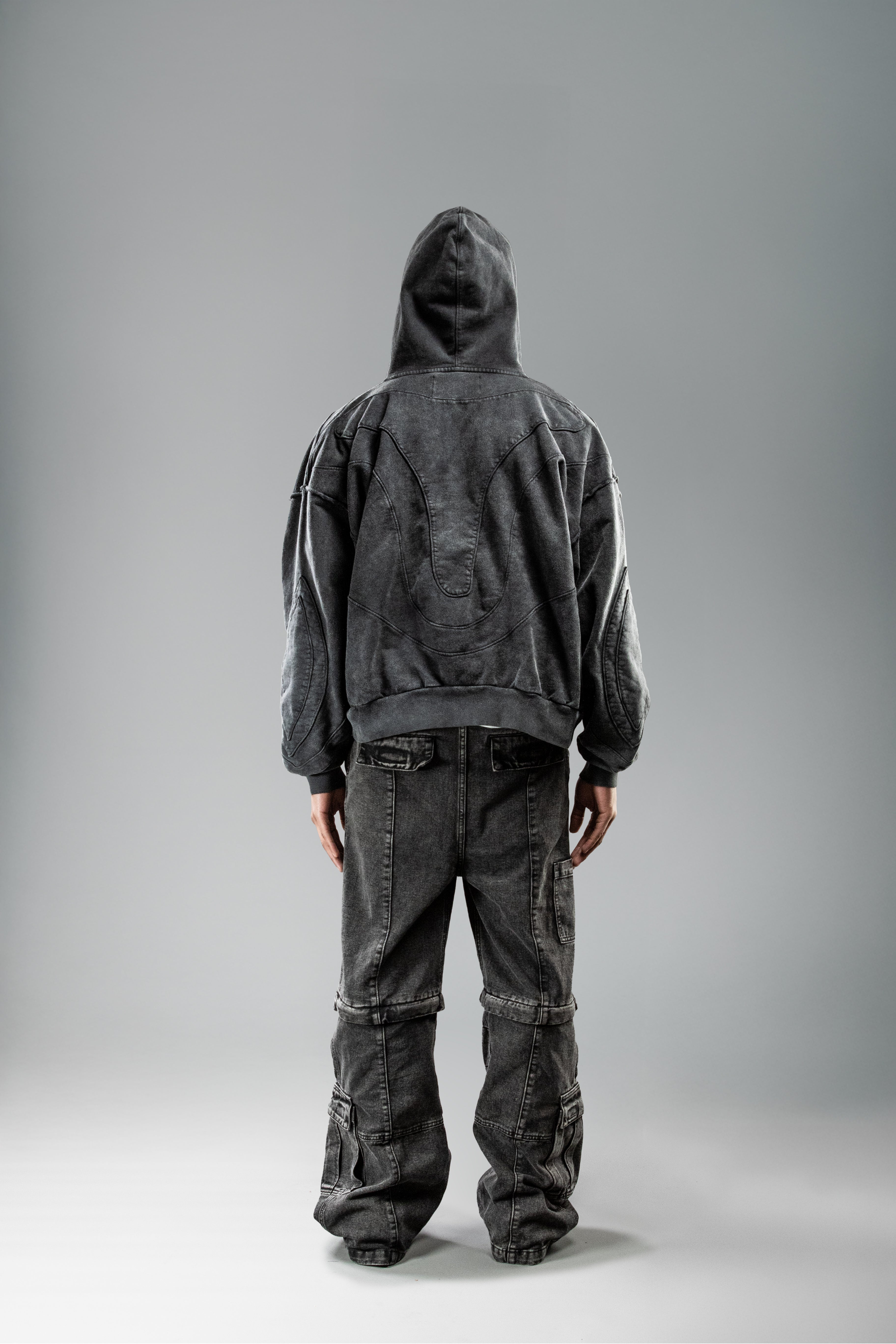 Getragene Rückenansicht des schwarzen Hoodies mit aufgesetzter Kapuze. Die Rückseite zeigt das geschwungene Nahtmuster in betonter Linienführung. Der Stoff wirkt leicht strukturiert und verwaschen.