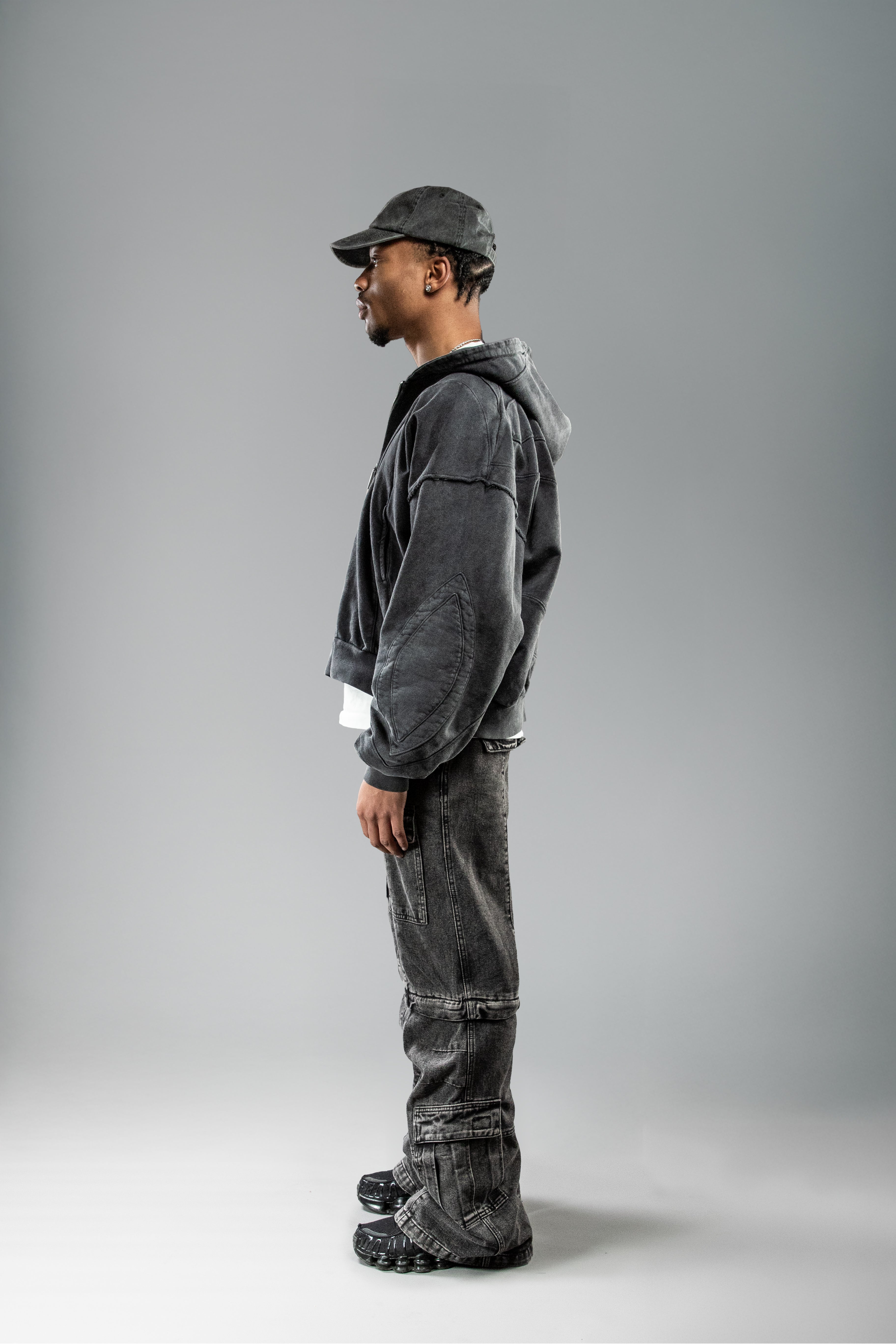 Getragene Seitenansicht des schwarzen Zip-Hoodies. Der Schnitt fällt gerade und kastig, die Kapuze ist locker auf den Rücken gelegt. Auffällig sind die elliptischen Nahtmuster entlang der Ärmelrundung.