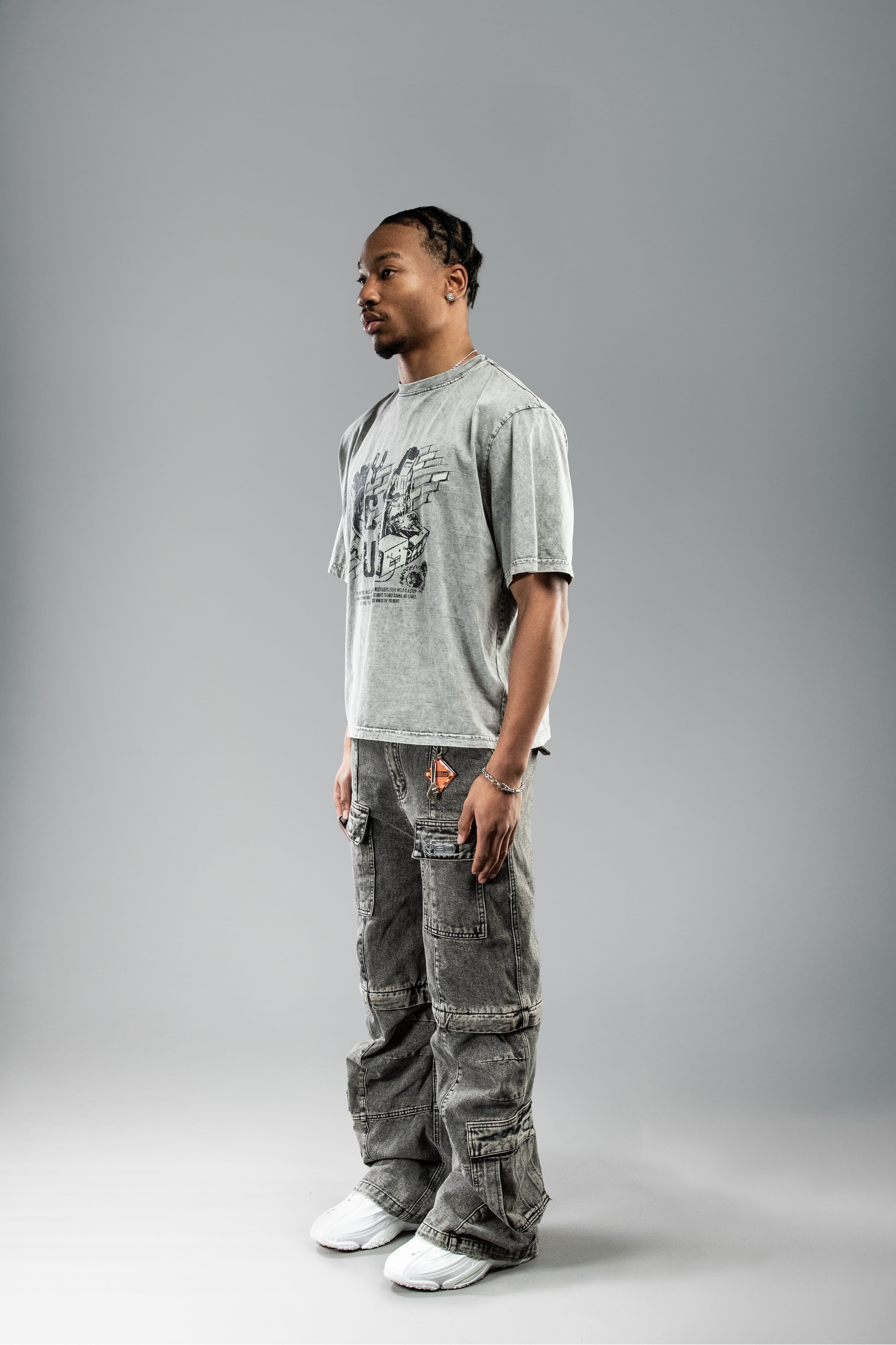 Getragenes Shirt von schräg vorn: graues Oversized-T-Shirt mit mechanischem Printmotiv, kombiniert mit dunkler Cargohose.