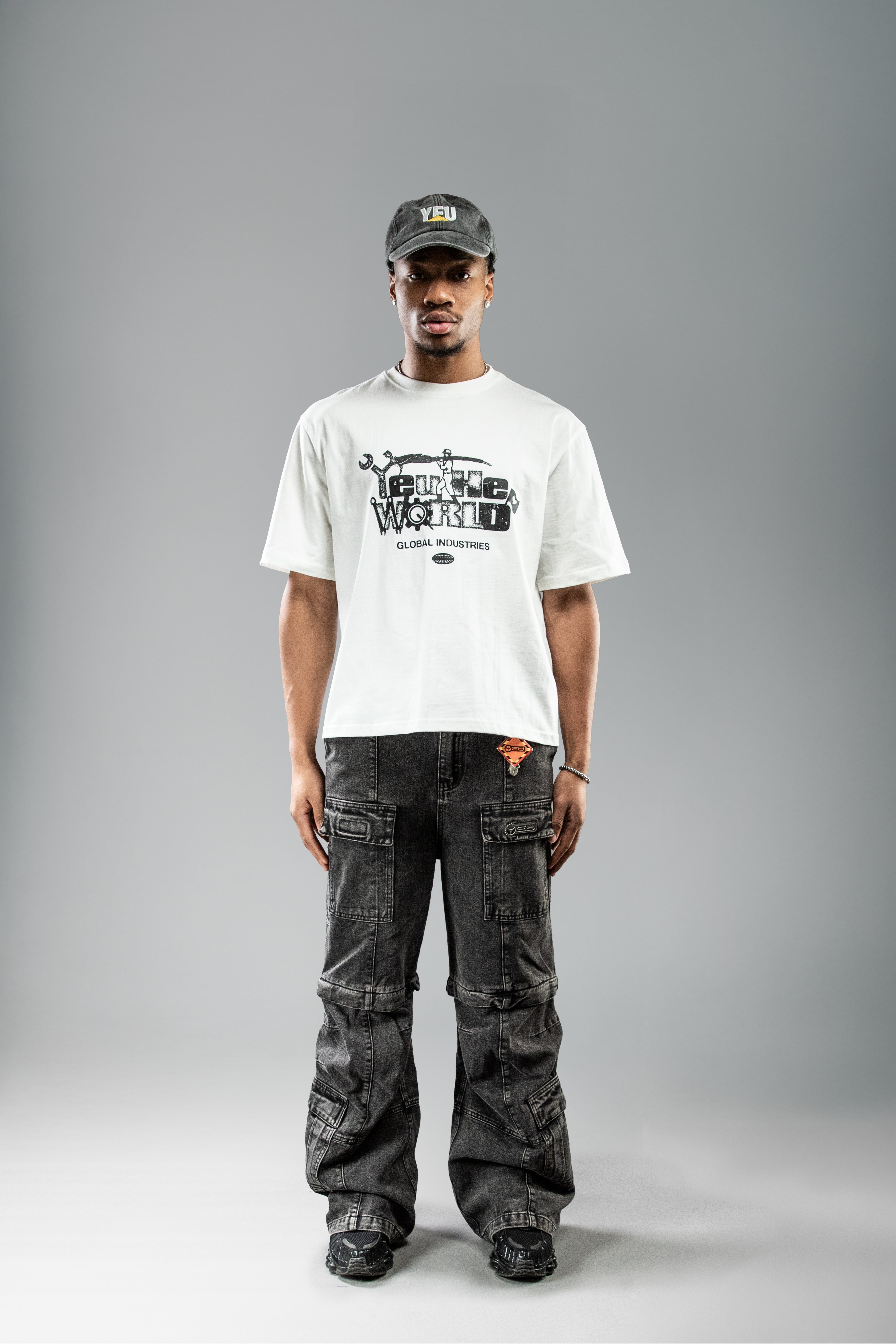 Weißes T-Shirt mit plakativem Print „FUTURE WORLD“ und Werkzeug-Motiven mittig auf der Brust. Darunter steht in kleinerer Schrift „GLOBAL INDUSTRIES“. Der Look ist gerade und locker geschnitten.
