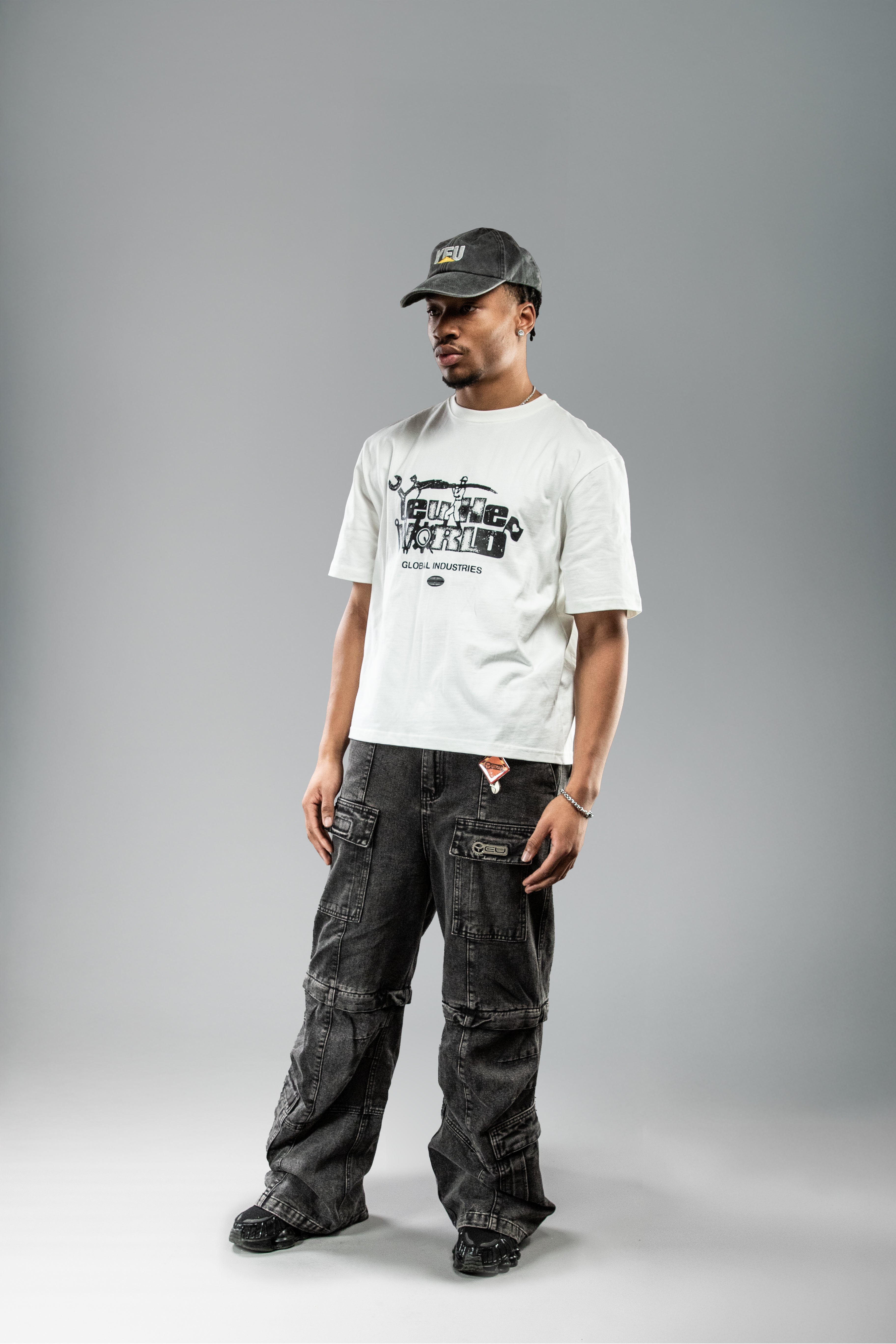 Weißes T-Shirt mit großem schwarzen Frontprint „FUTURE WORLD“ samt Werkzeugillustrationen, getragen in leichter Drehung. Dazu: dunkle Cargohose mit Label-Patch und Cap.