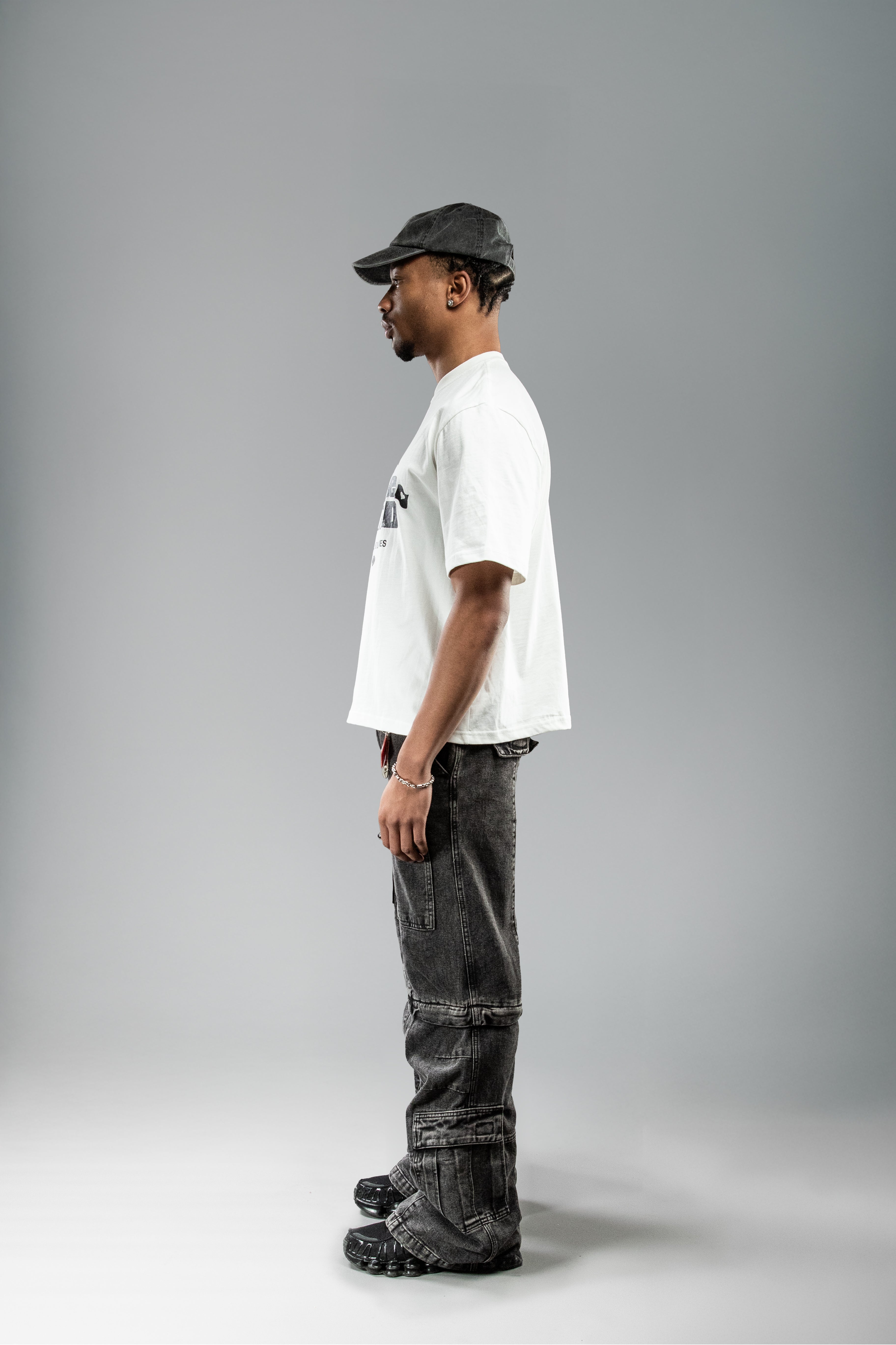 Getragenes weißes T-Shirt in Seitenansicht. Der schwarze Frontprint ist nur angeschnitten sichtbar. Kombiniert mit schwarzer Cap und grauer Cargo-Hose.